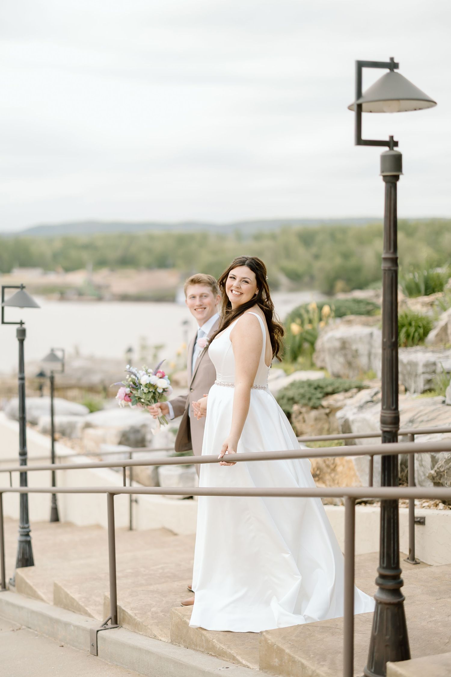 Two people in white dresses sit on a stone wall overlooking a scenic desert landscape with decorative lampposts.
