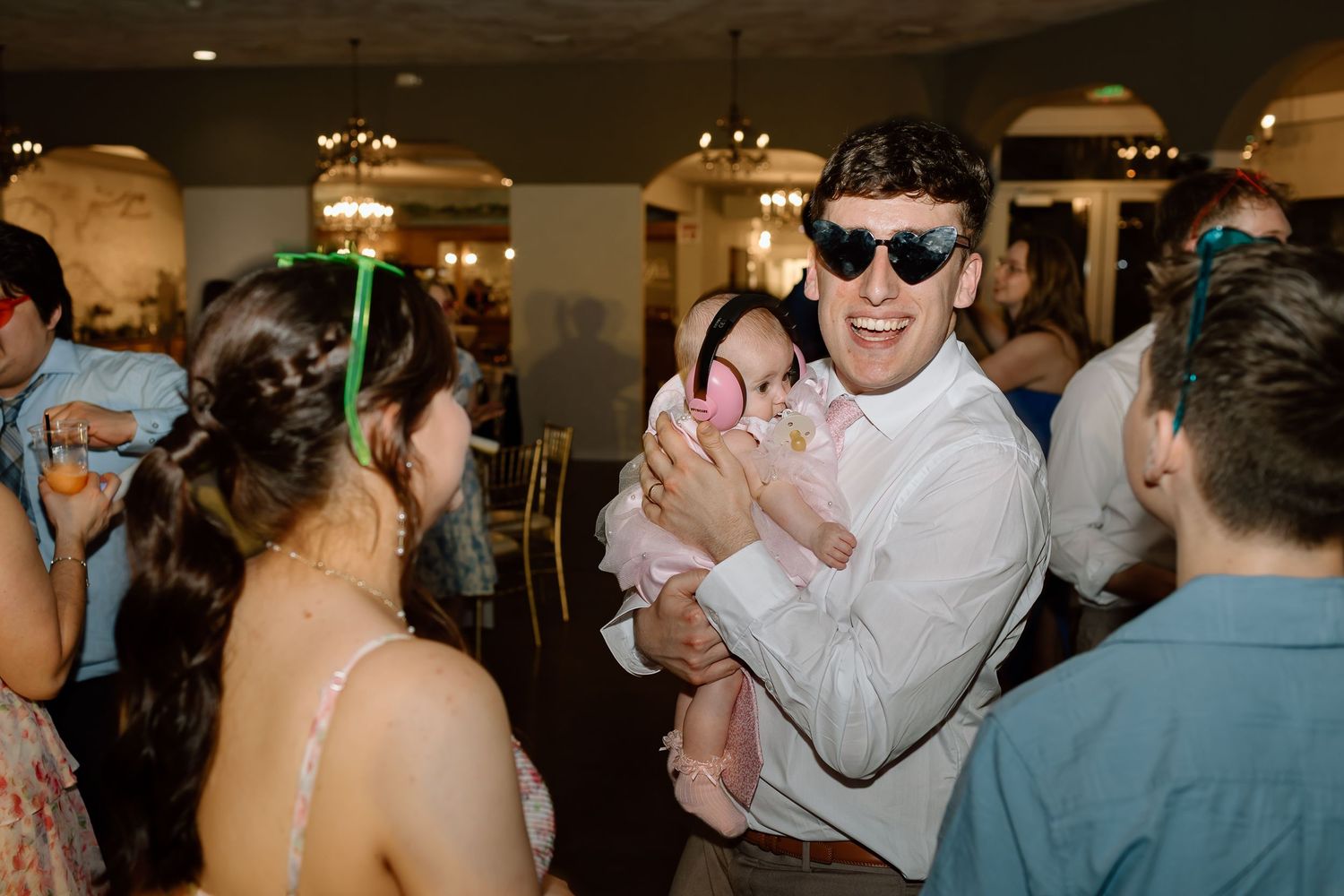 Wedding guests dance and celebrate on a crowded dance floor at an indoor reception venue.