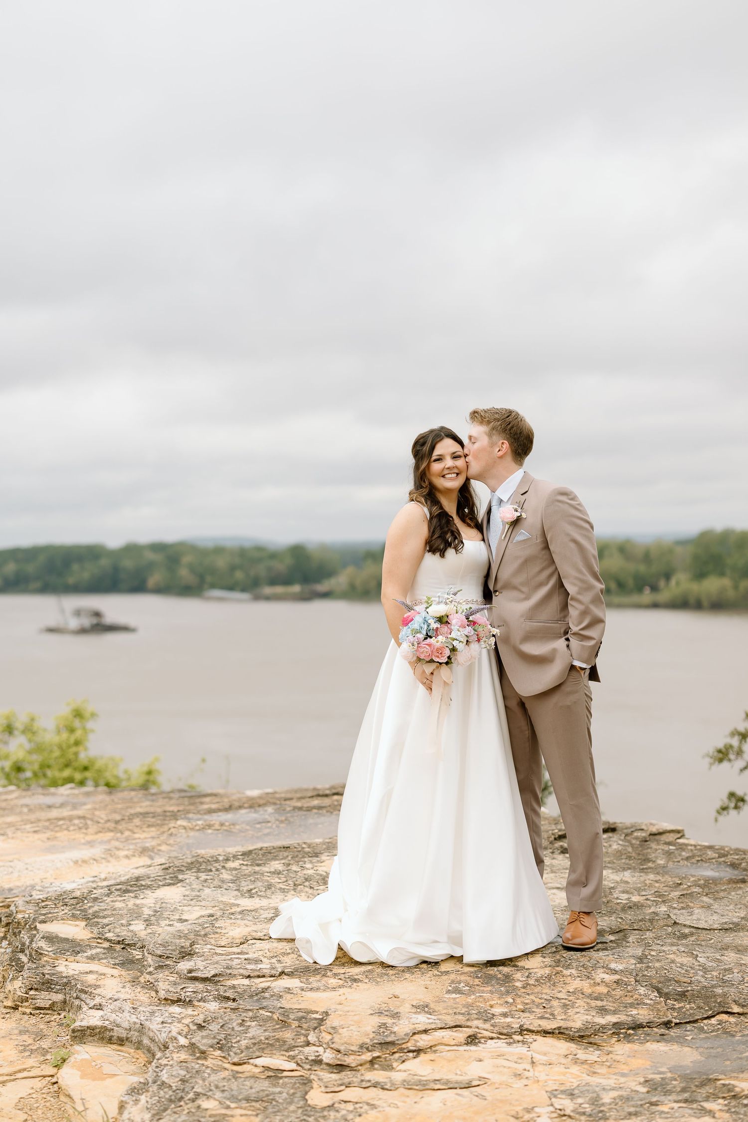 A couple shares an intimate moment on a rocky cliff overlooking a serene lake on their wedding day.