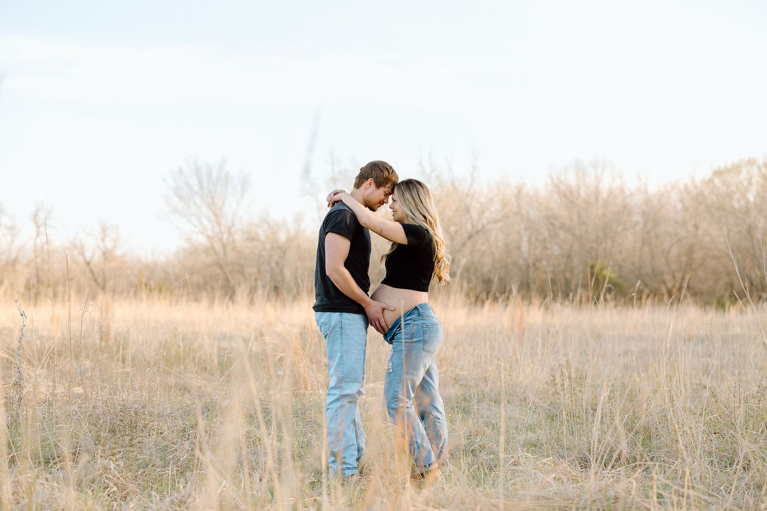 A couple embraces while standing in a sunlit field of tall grass during golden hour.