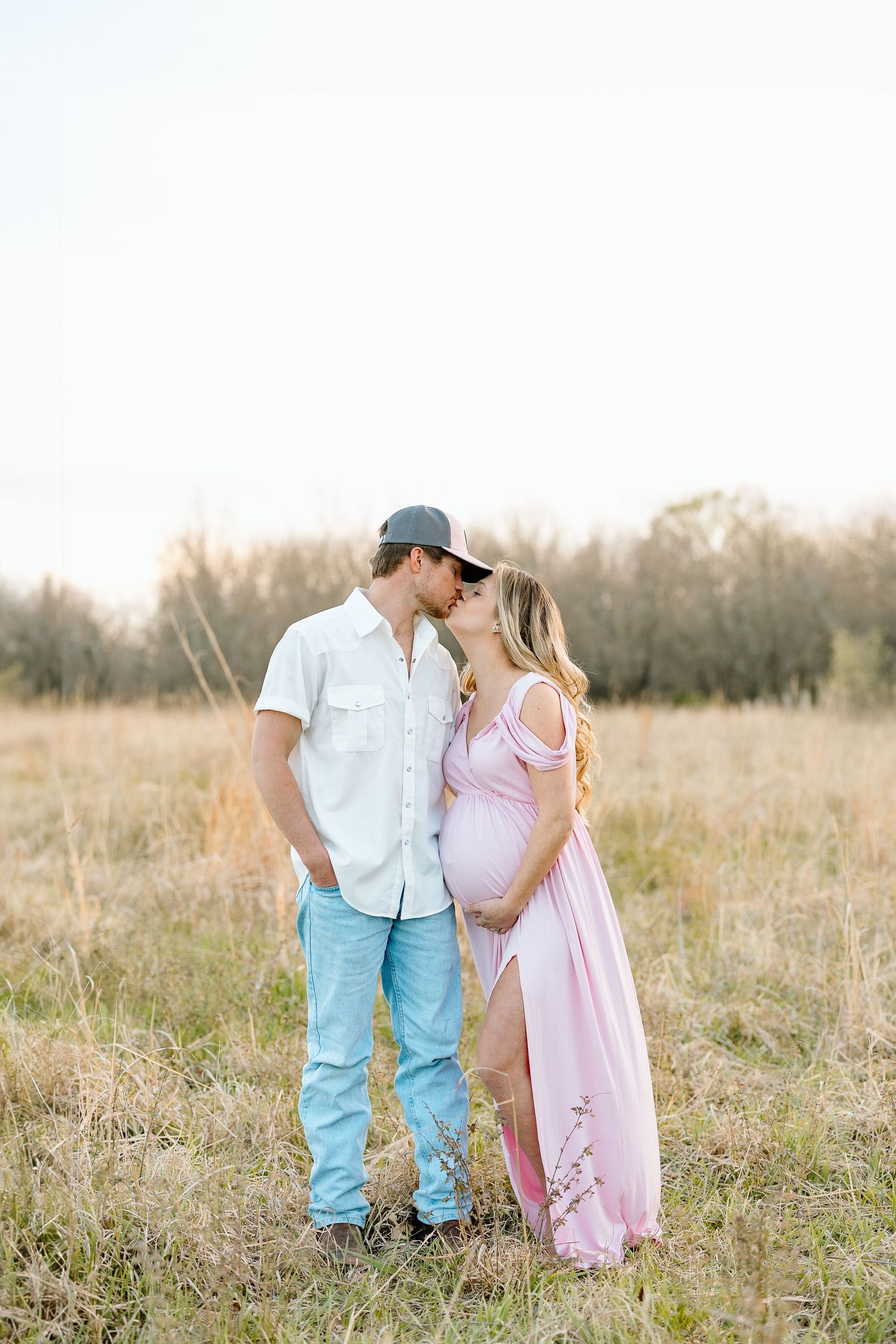 A romantic couple in casual attire stands together in a sunlit meadow with tall grass at golden hour.