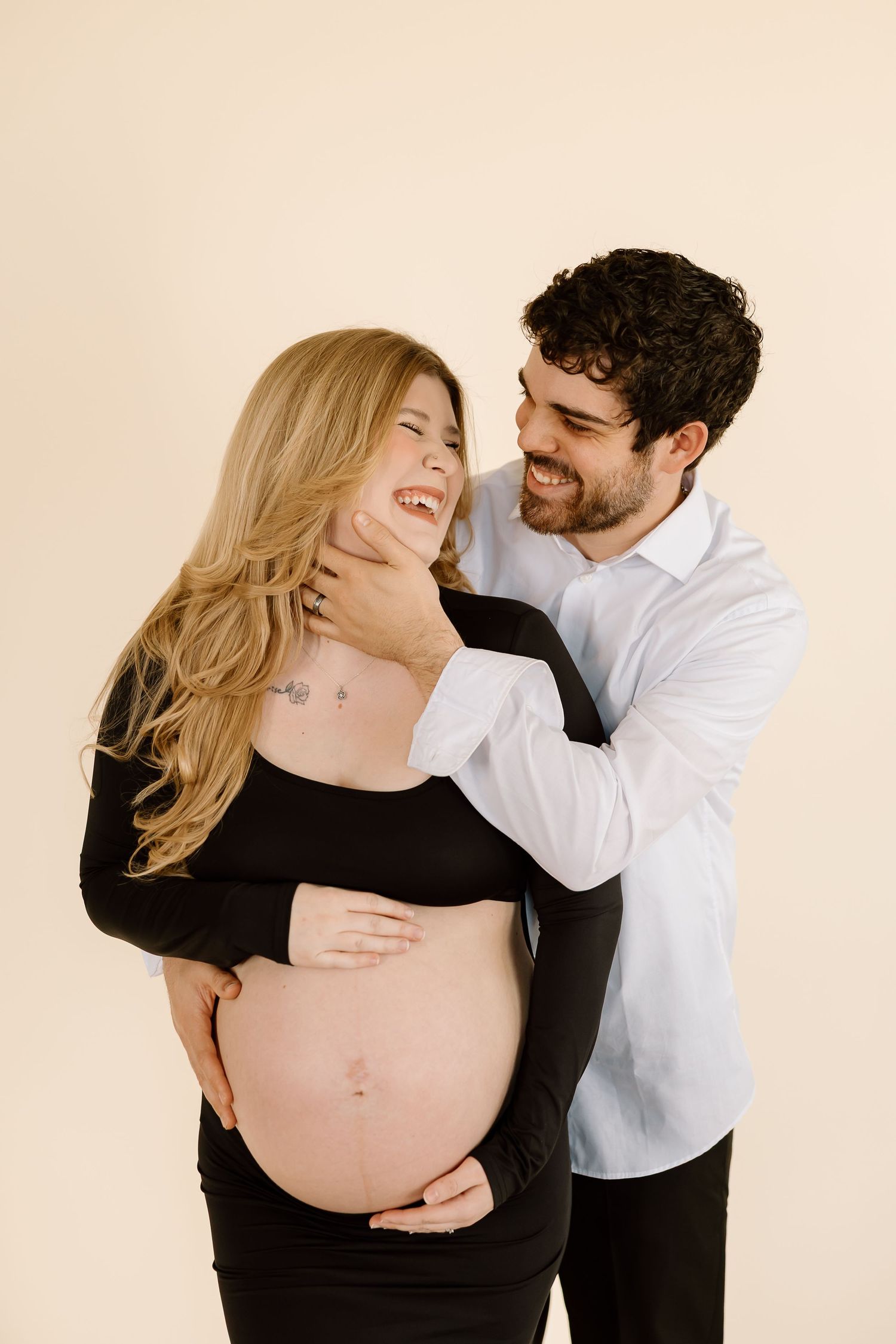 A couple shares intimate moments during a maternity photoshoot showing a pregnant woman in black dress and partner in white shirt.