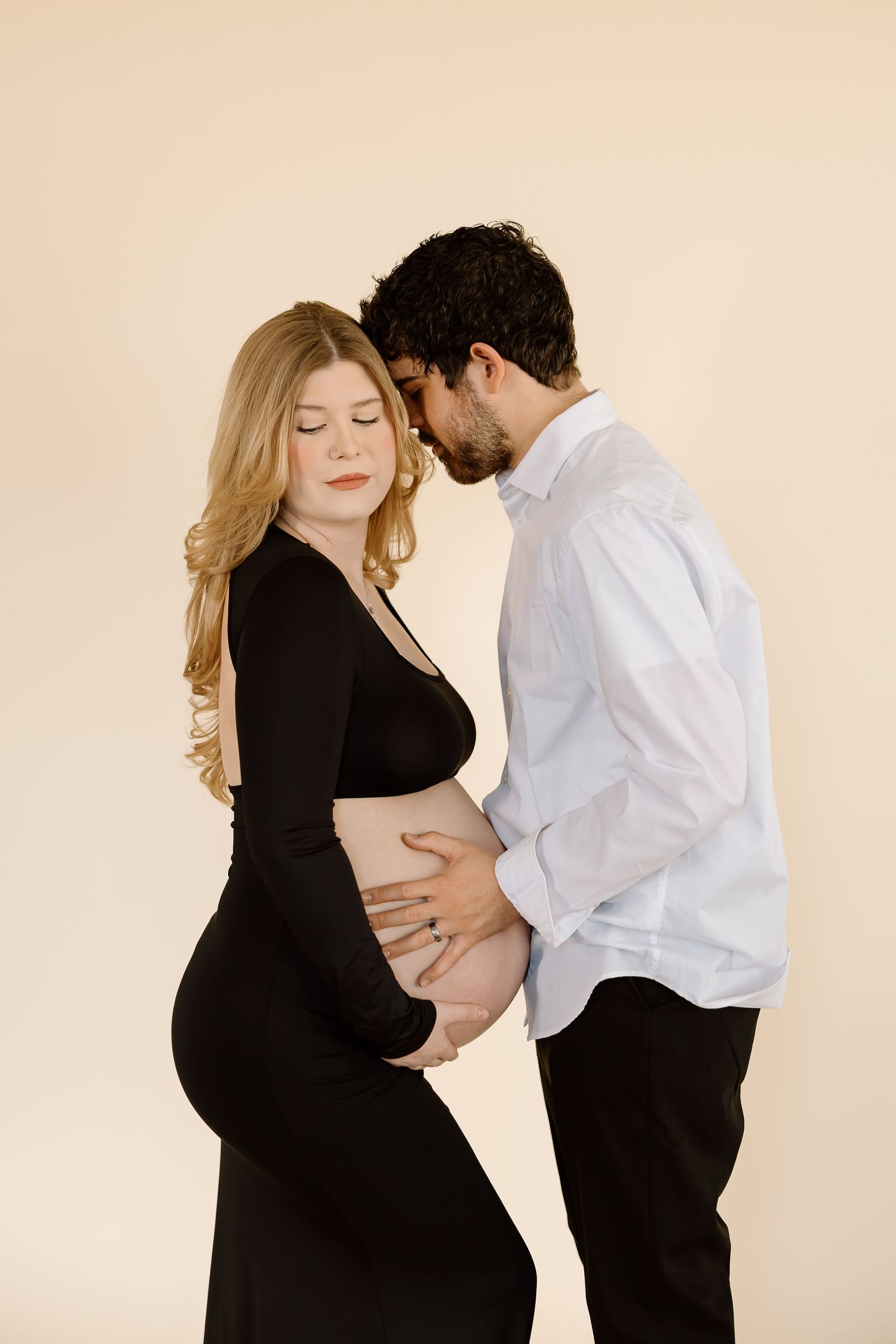 Intimate maternity photo of couple in black and white outfits embracing against beige backdrop.
