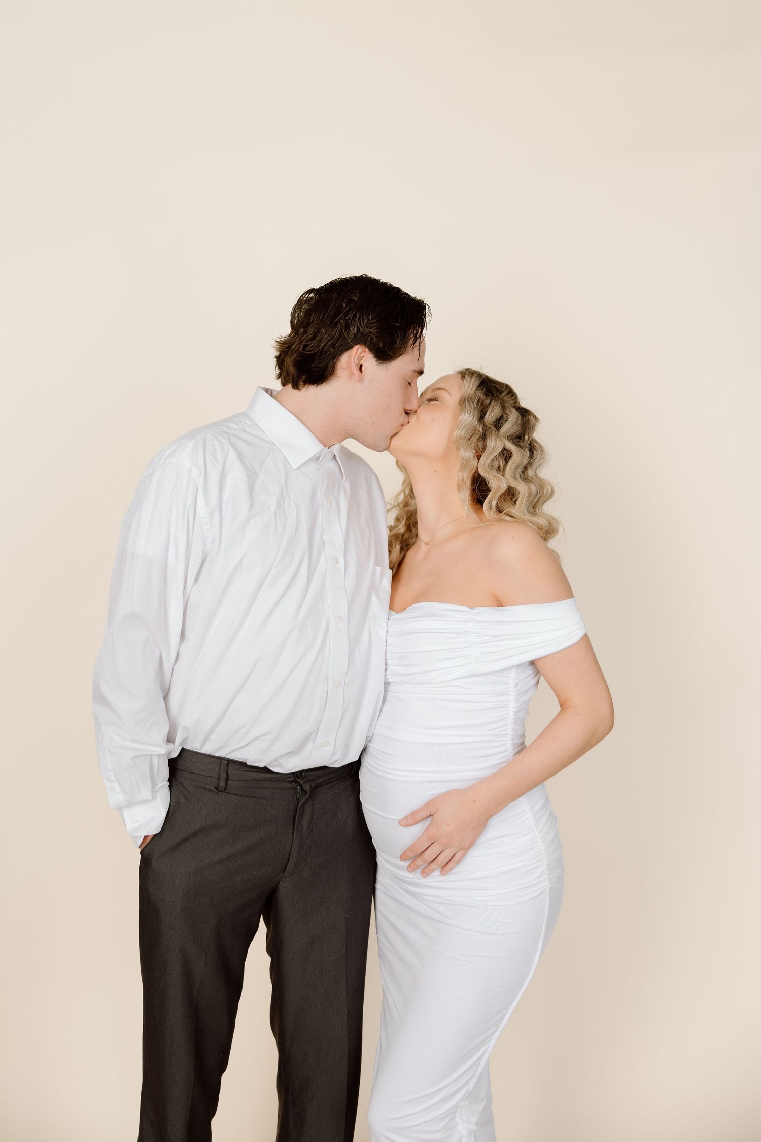 A couple in formal attire share an intimate kiss against a neutral background.