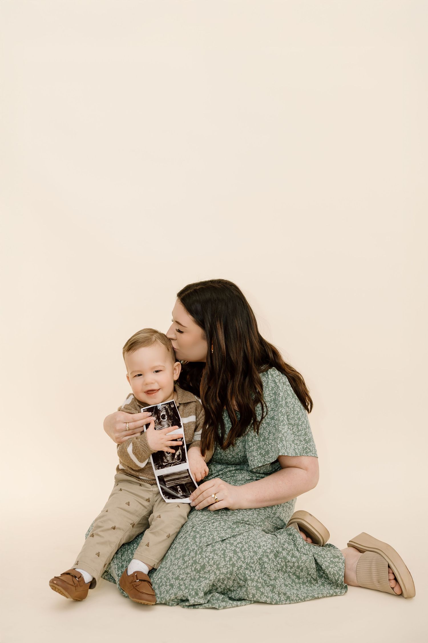 A woman in a green floral dress sits on a cream background with a small child in neutral colored clothing.