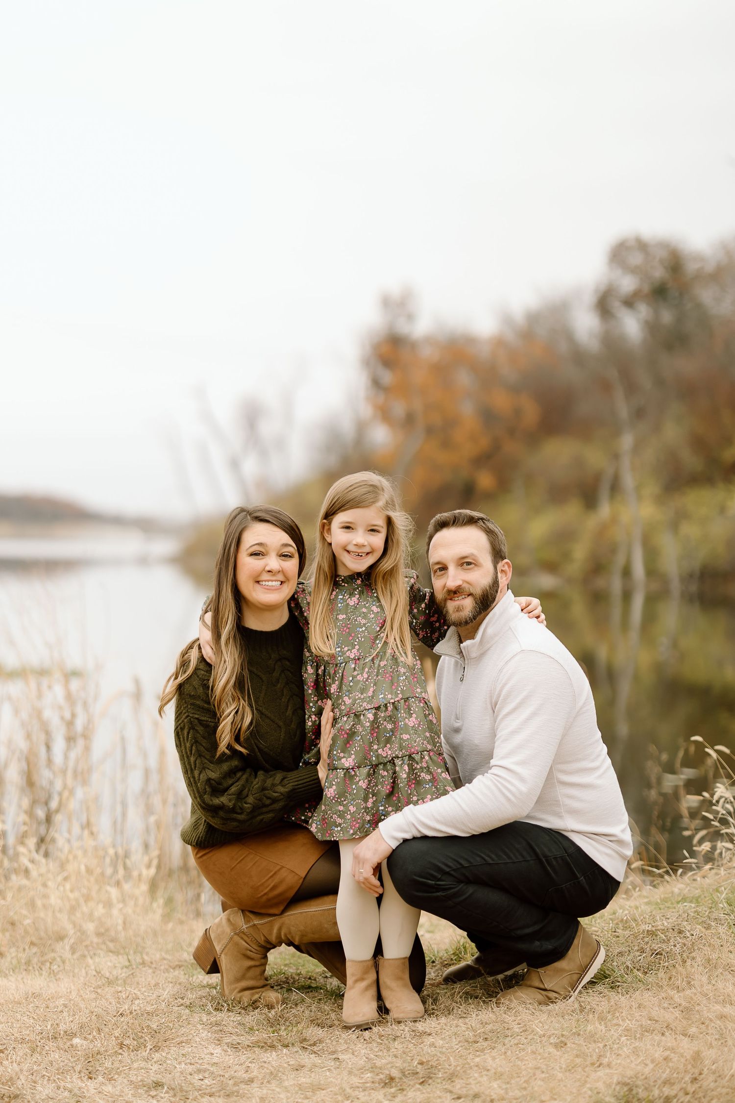 Family poses together on autumn day near lake with fall foliage in background.