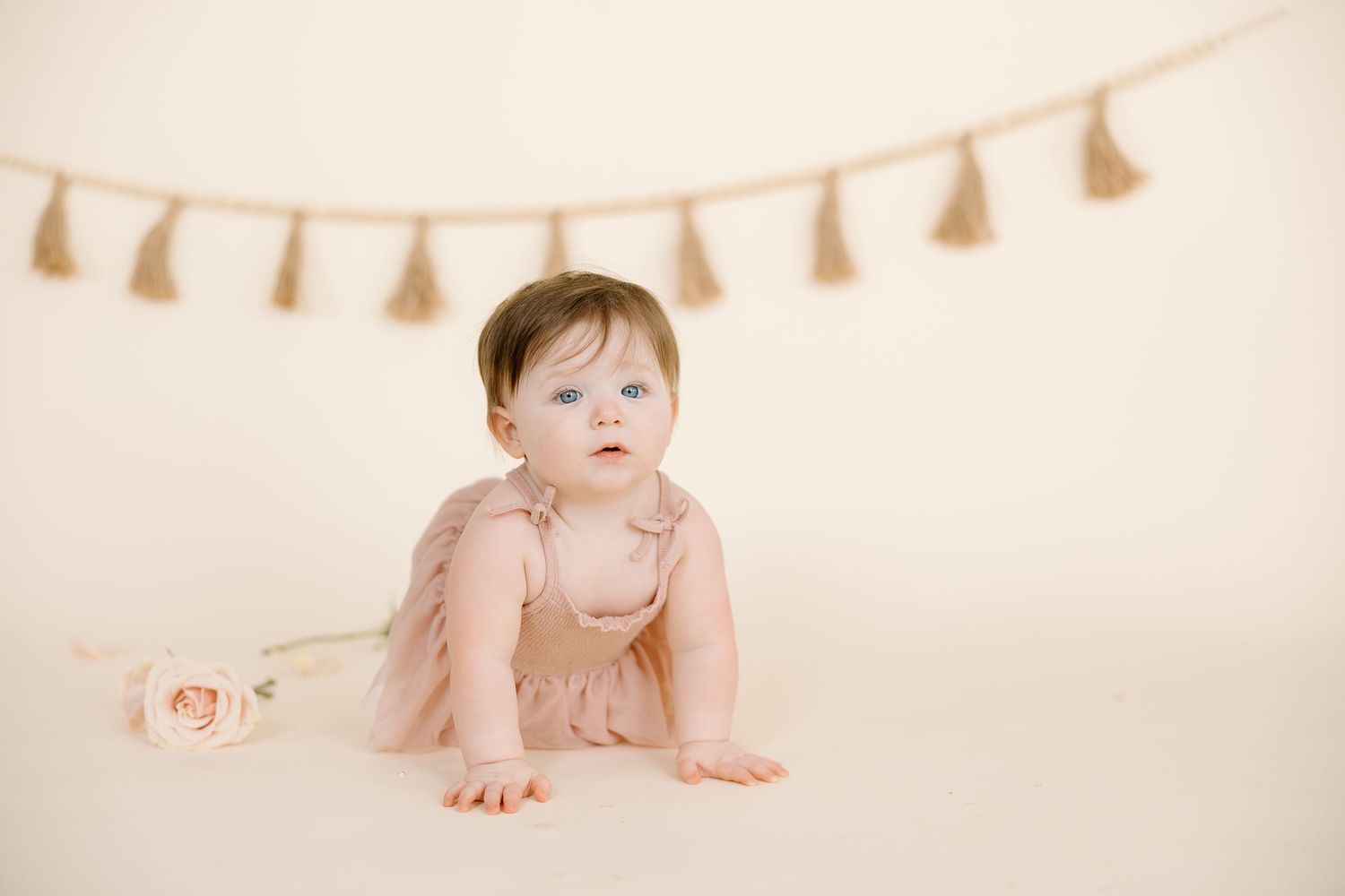 A baby in pink clothing poses beneath a beige tassel garland with roses.