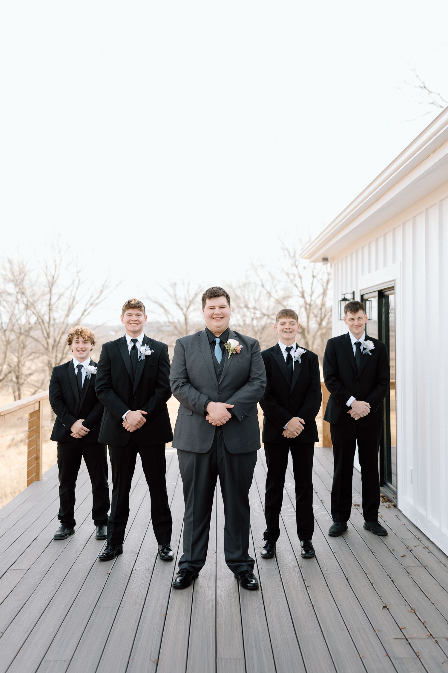 Groomsmen in black suits pose formally on wooden deck.