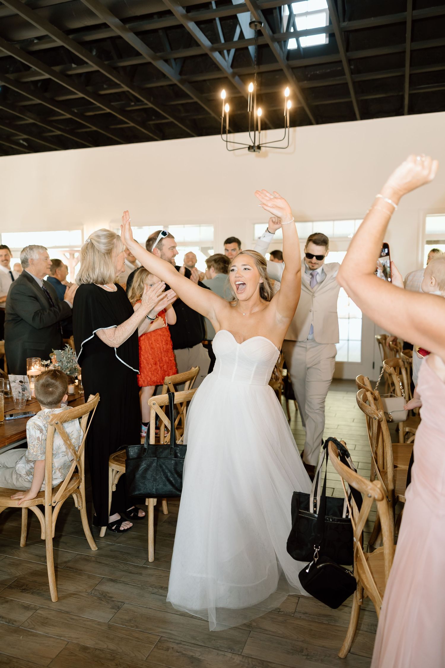 Reception celebration with guests dancing and raising arms in rustic indoor venue with wooden ceiling.