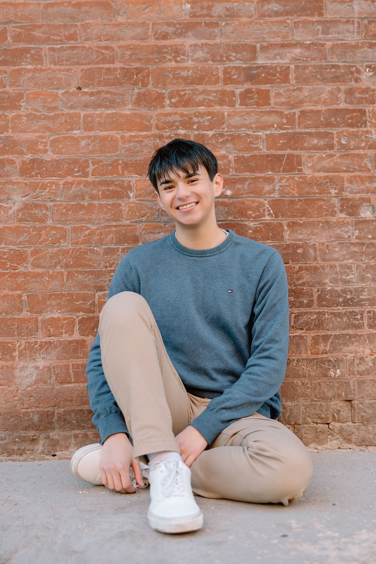 Relaxed portrait sitting cross-legged against rustic brick background.