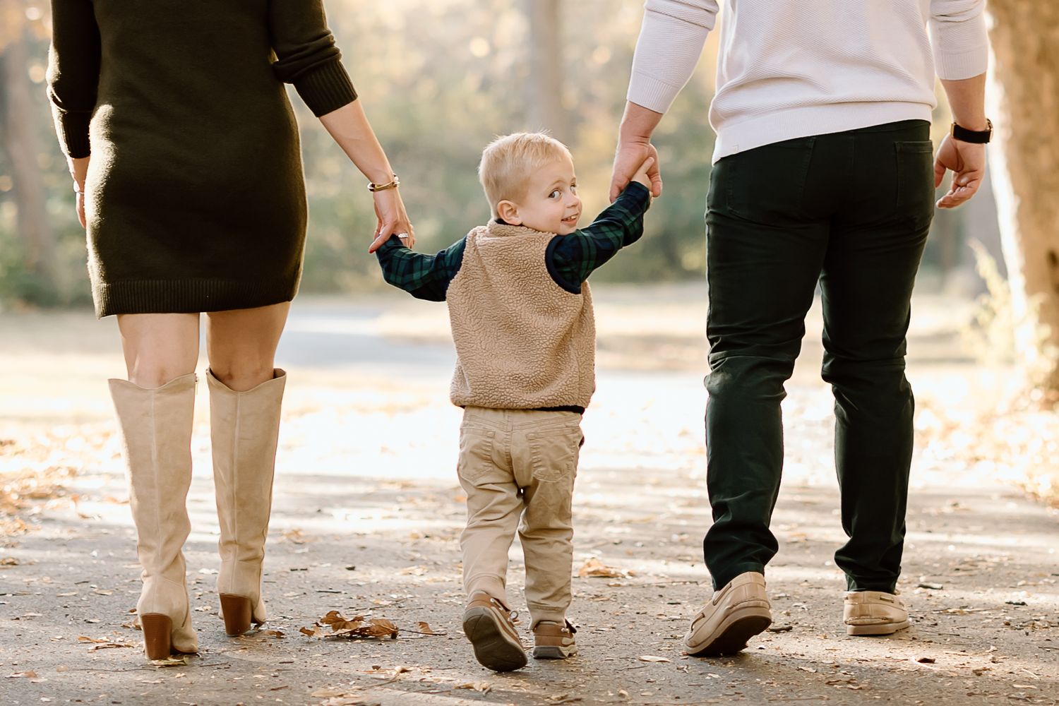 A toddler holds hands with two adults while walking on a leaf-covered path during autumn.
