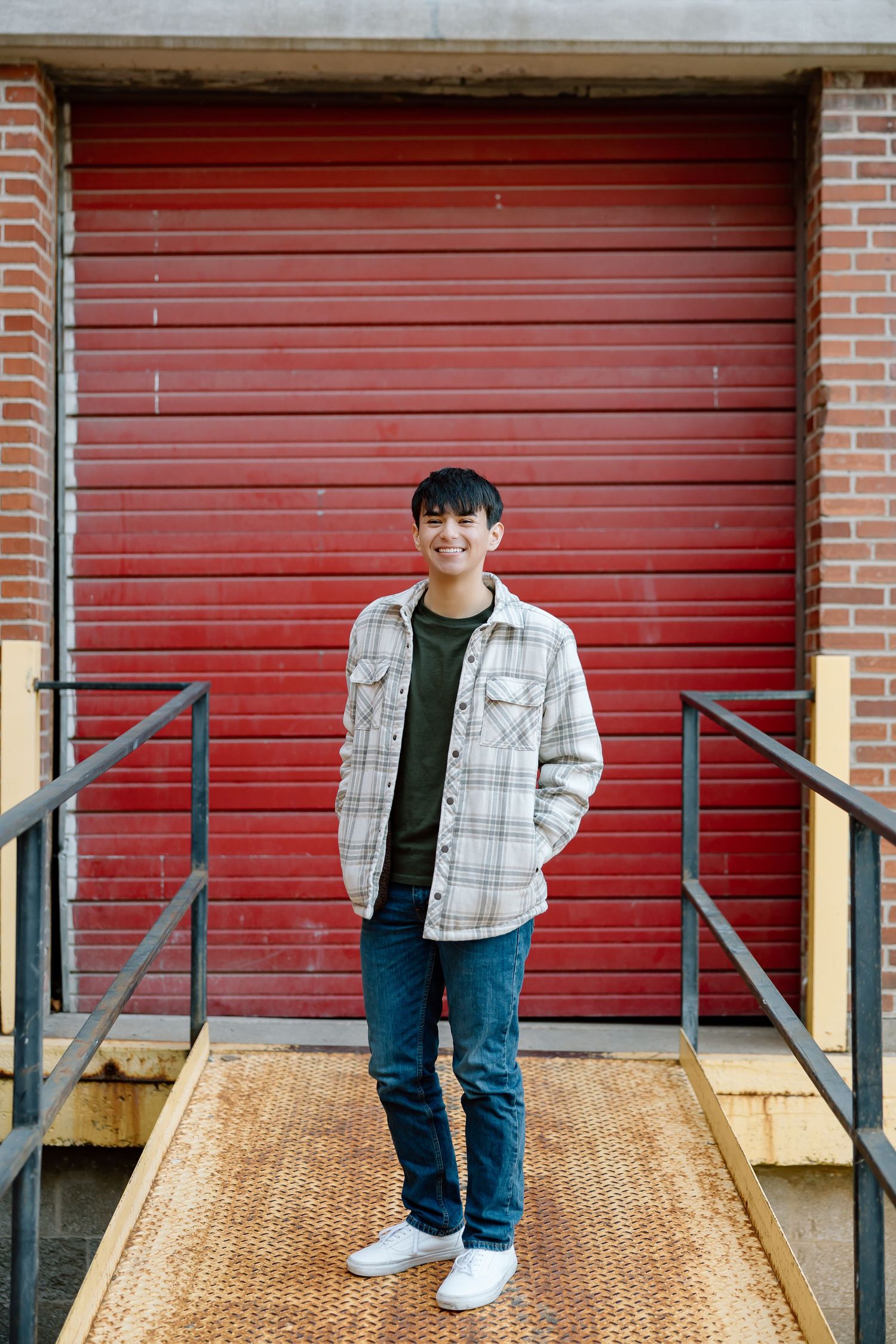 Person in plaid shirt and jeans standing in front of red garage door with railings.