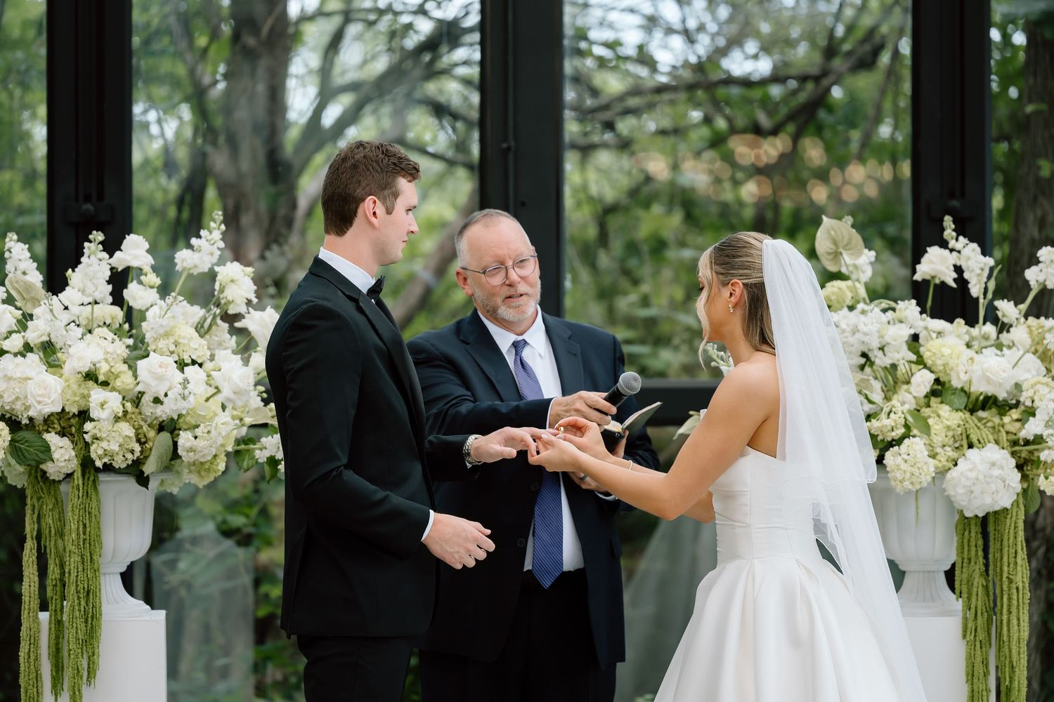 Groom places wedding ring on bride's finger during elegant indoor ceremony with white floral arrangements.