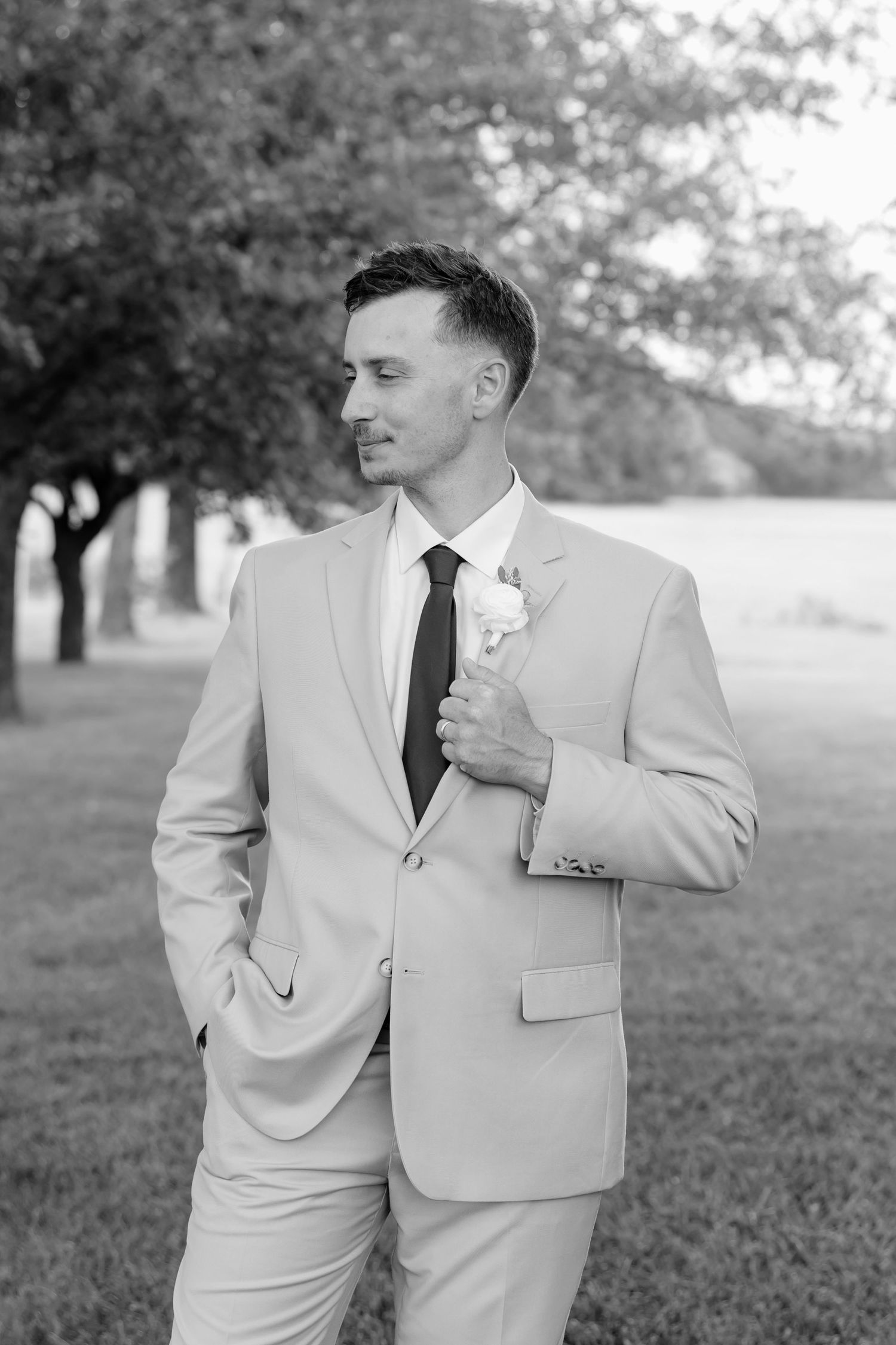 Groom in light suit with boutonniere poses outdoors among trees in elegant black and white wedding portrait.