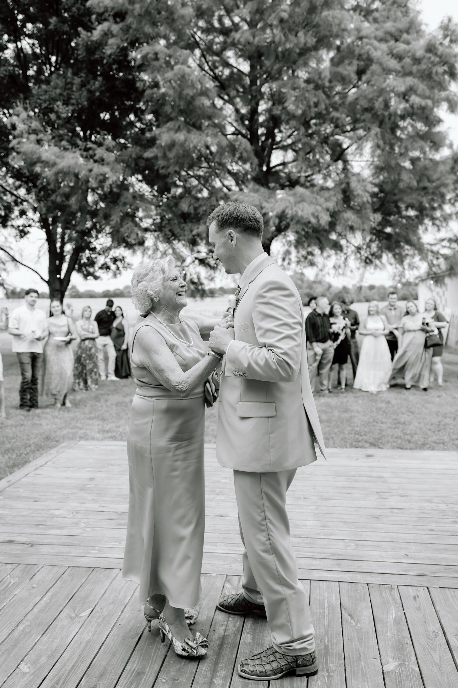 Black and white photo of a groom dancing with his mother on a wooden deck at an outdoor wedding reception.