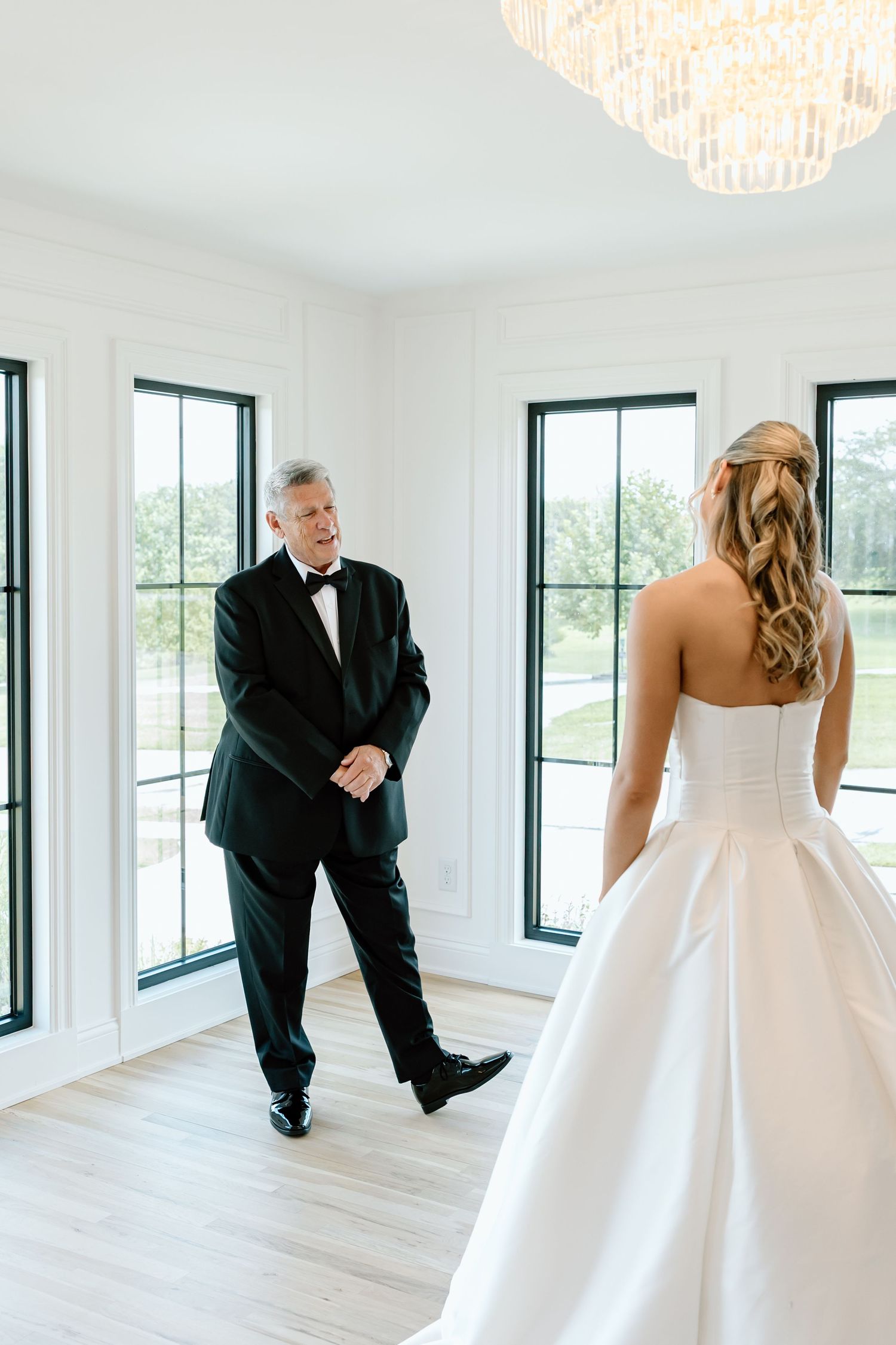 Father sees bride in wedding gown for first time during emotional first look in bright white room.