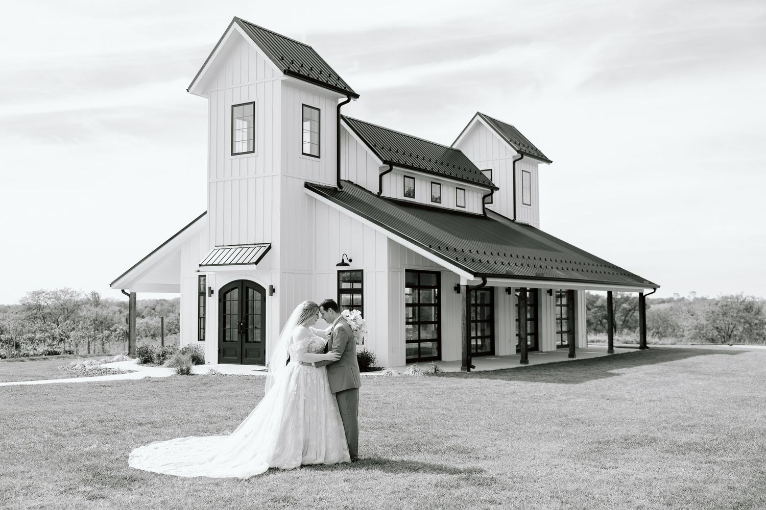 A black and white photo of a couple in wedding attire standing in front of a rustic white barn venue.