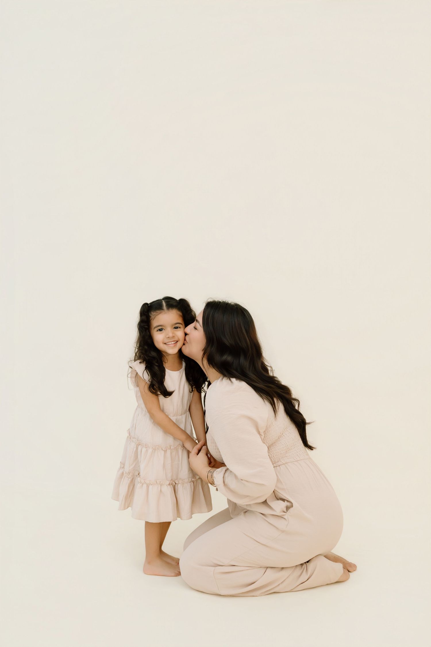 A tender moment captured against a white backdrop as two figures in neutral-toned dresses share an affectionate pose.