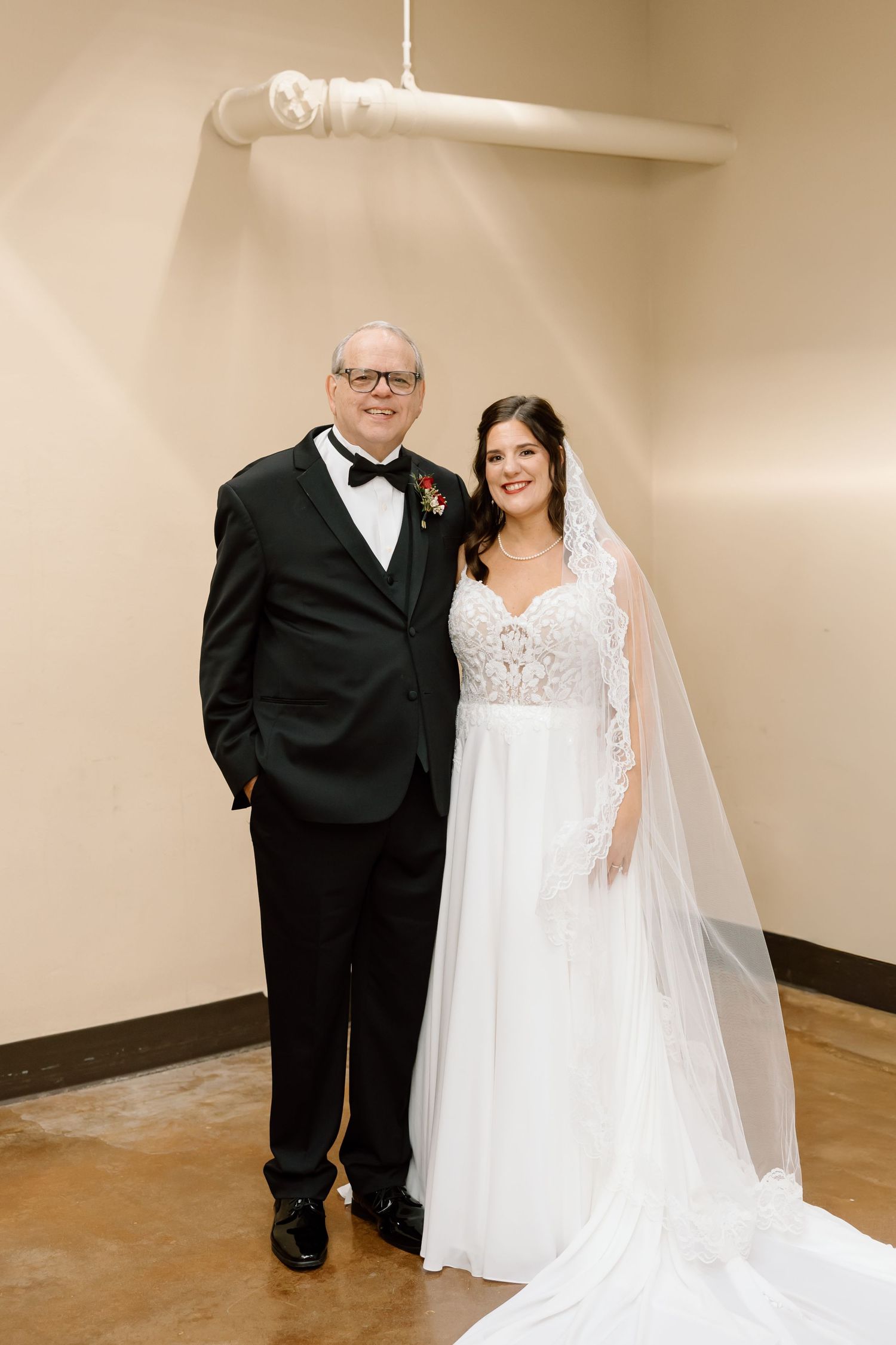 A wedding couple poses in formal attire against a beige wall, with the bride wearing a flowing white lace gown and veil.
