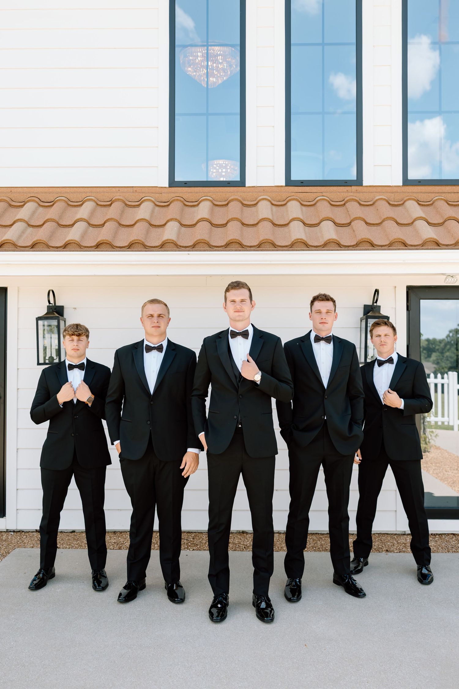 Five groomsmen in black tuxedos with bow ties pose confidently outside a white building with terracotta roof tiles.