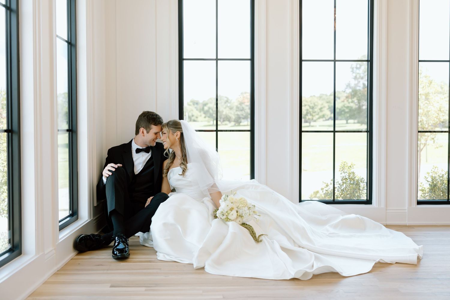Bride and groom sharing intimate moment seated on floor by large windows in elegant wedding venue.