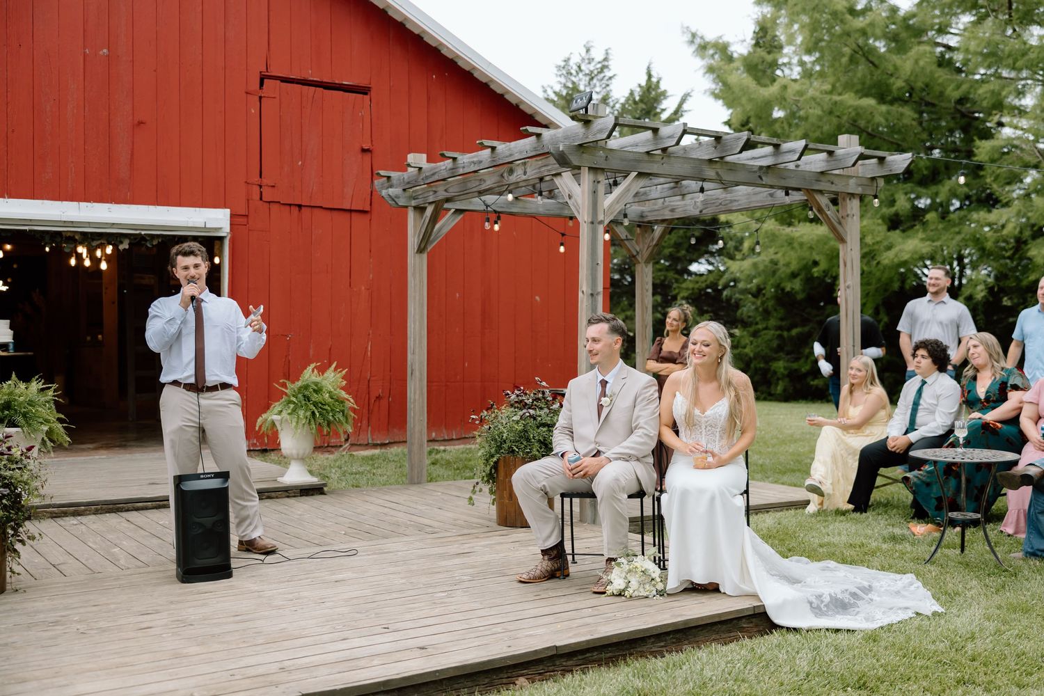 Groom and bride seated outdoors at rustic barn wedding venue while guest delivers speech on wooden deck.