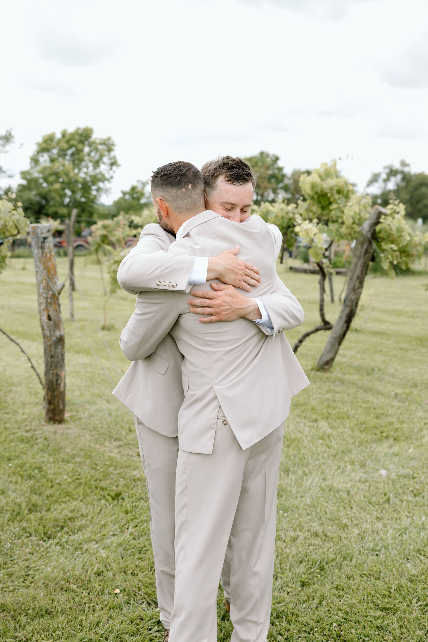Two grooms in matching beige suits share an emotional hug outdoors at a vineyard wedding ceremony.