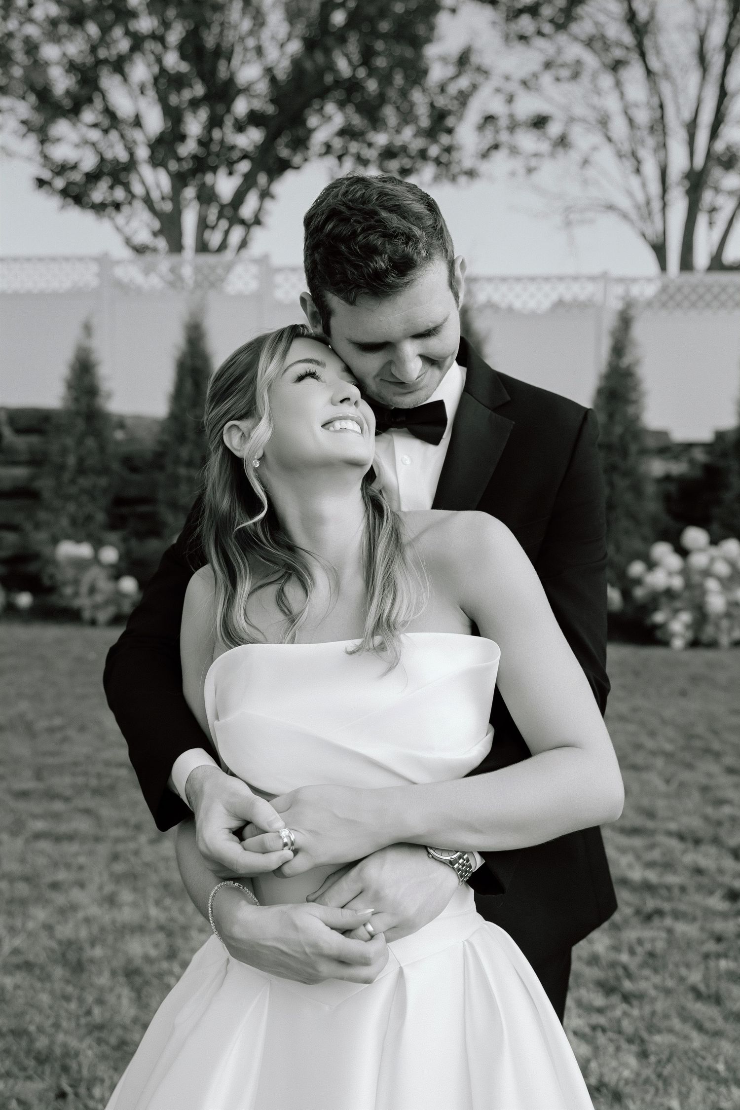Black and white photo of a bride and groom embracing outdoors, smiling joyfully in wedding attire.