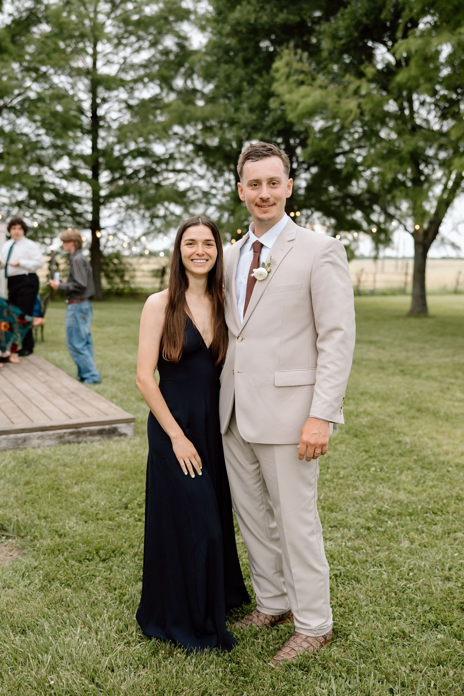 A couple poses outdoors at a wedding, woman in black dress and man in beige suit with floral boutonniere on green lawn.