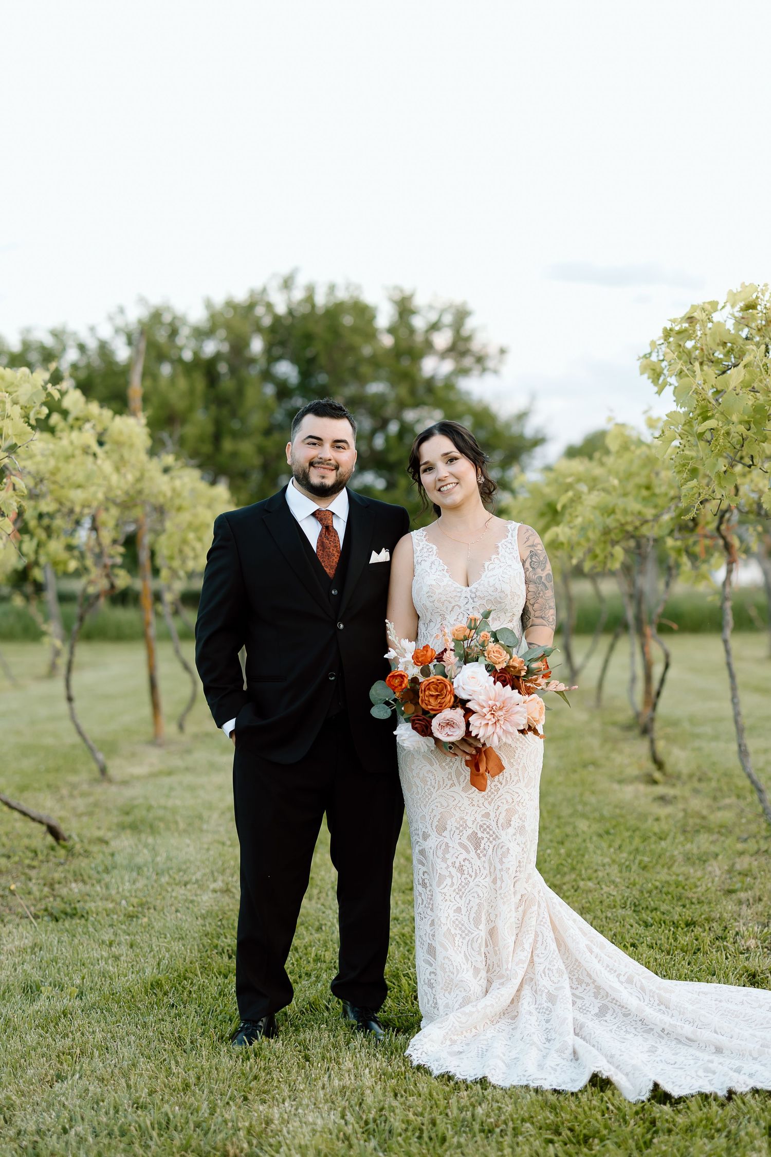 Couple stands together in a vineyard garden for their wedding portrait with bride holding orange and white bouquet.