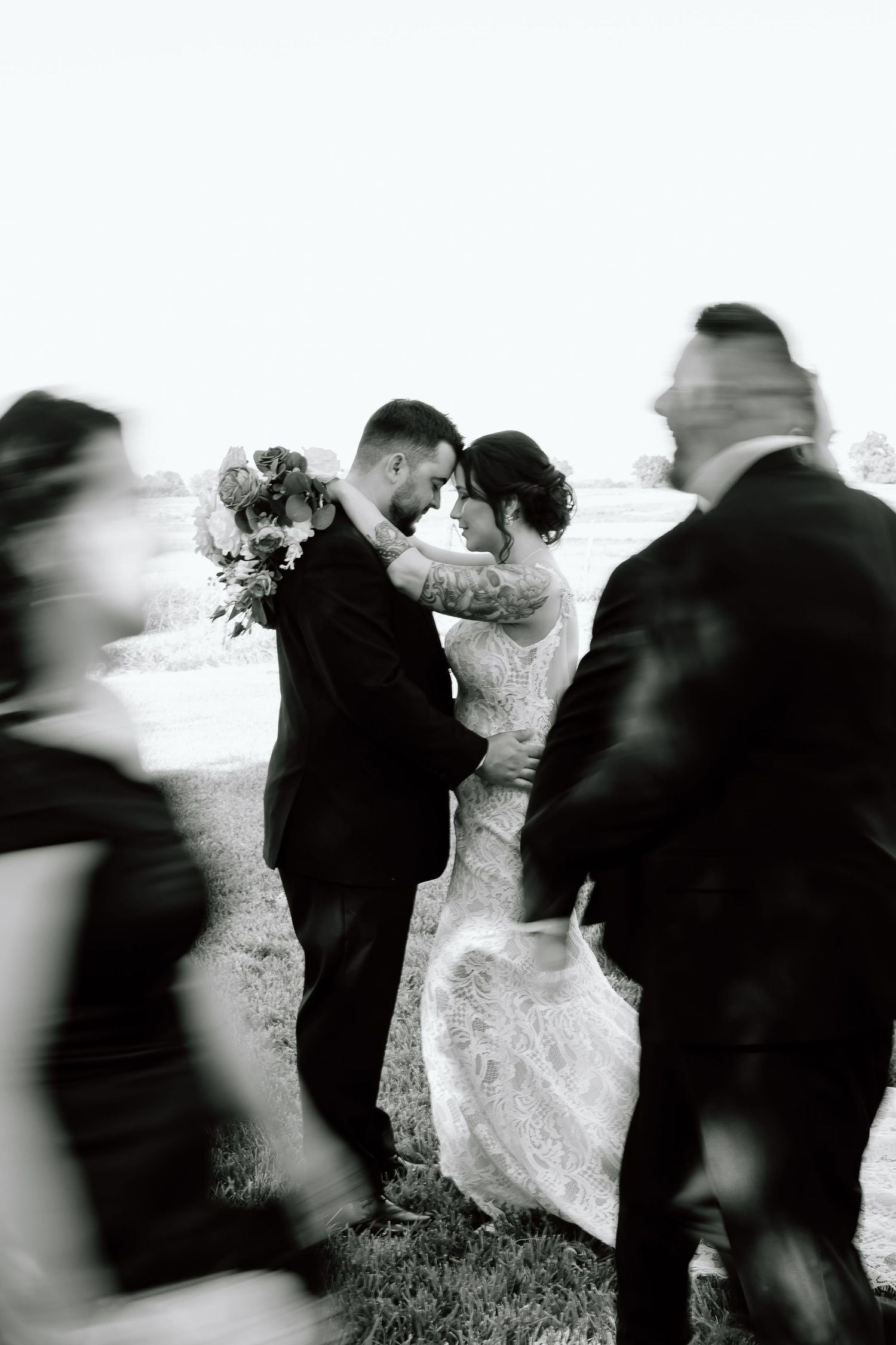 Black and white artistic wedding photo with blurred motion of people moving around a couple sharing an intimate moment outdoors.