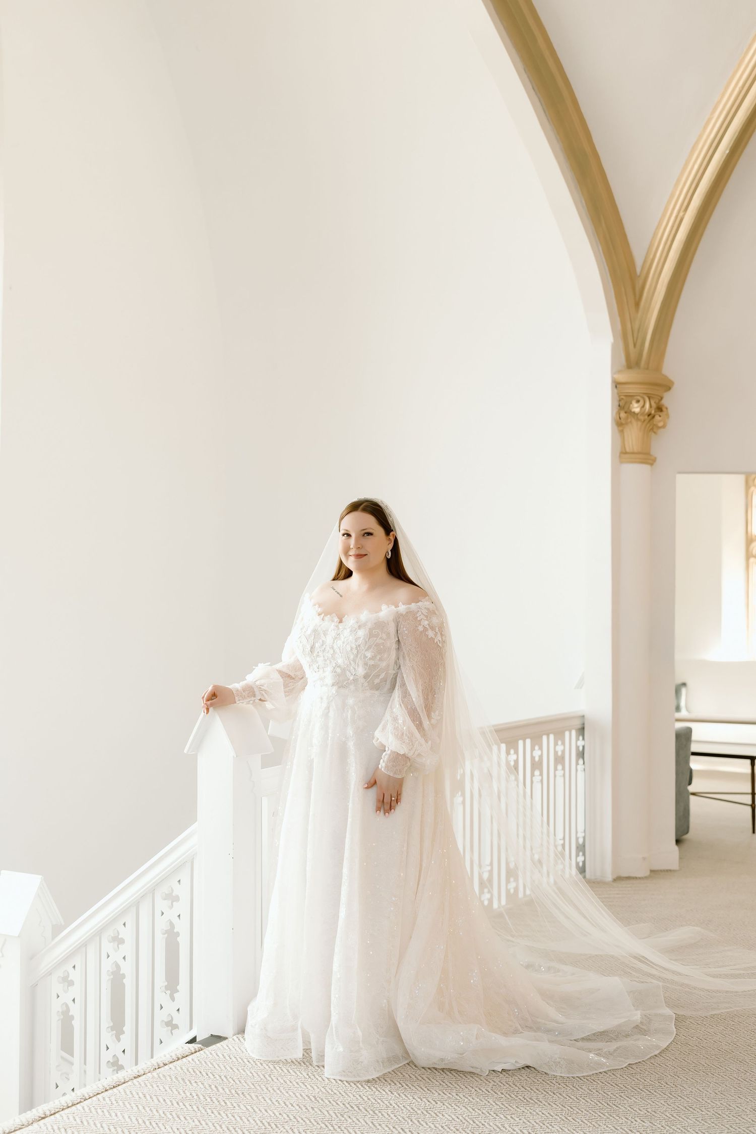 Bride in white lace off-shoulder gown with veil stands on elegant staircase in bright venue with arched ceilings.