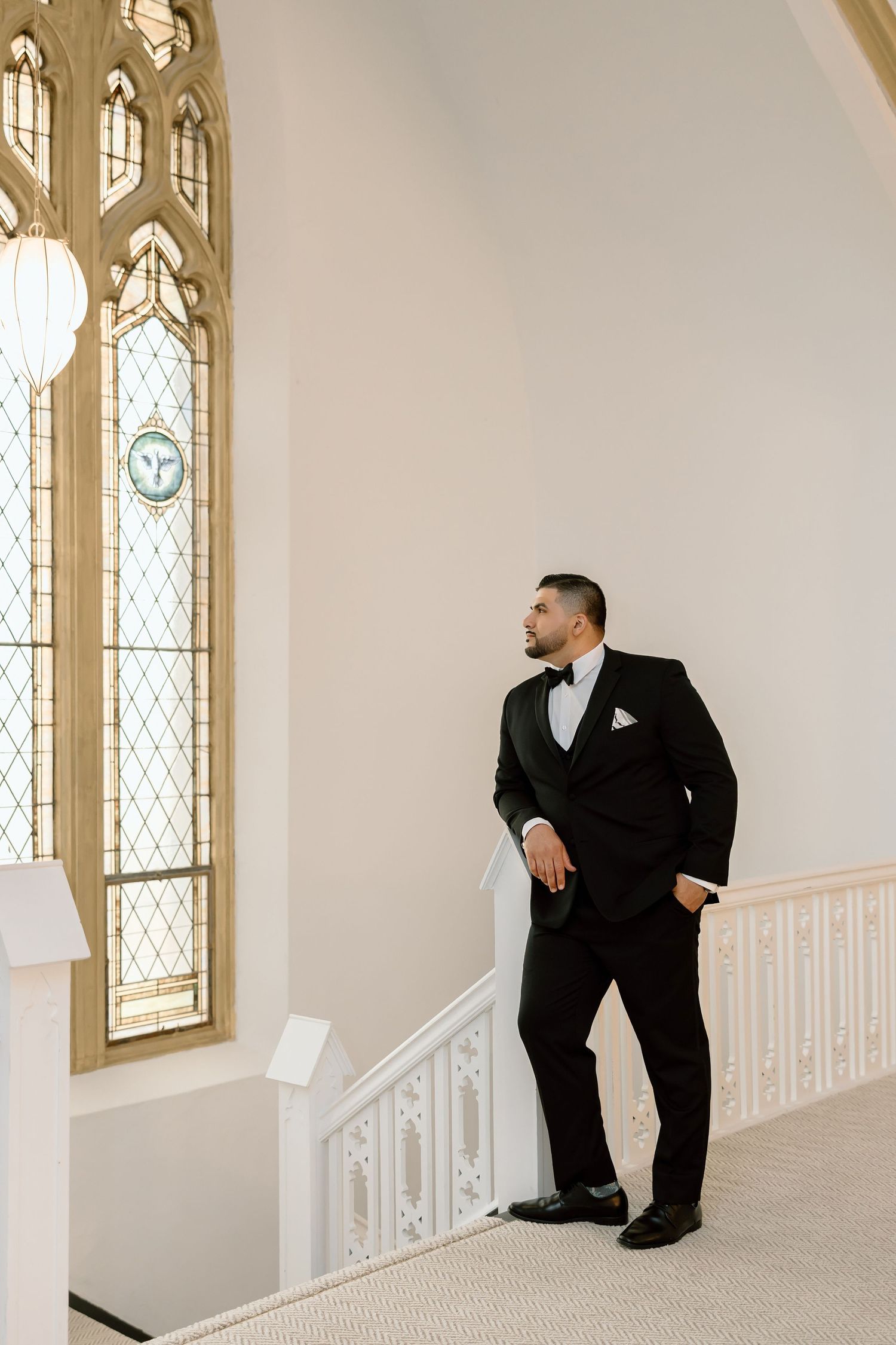 Groom in black tuxedo with bow tie stands by ornate stained glass window in elegant white chapel.