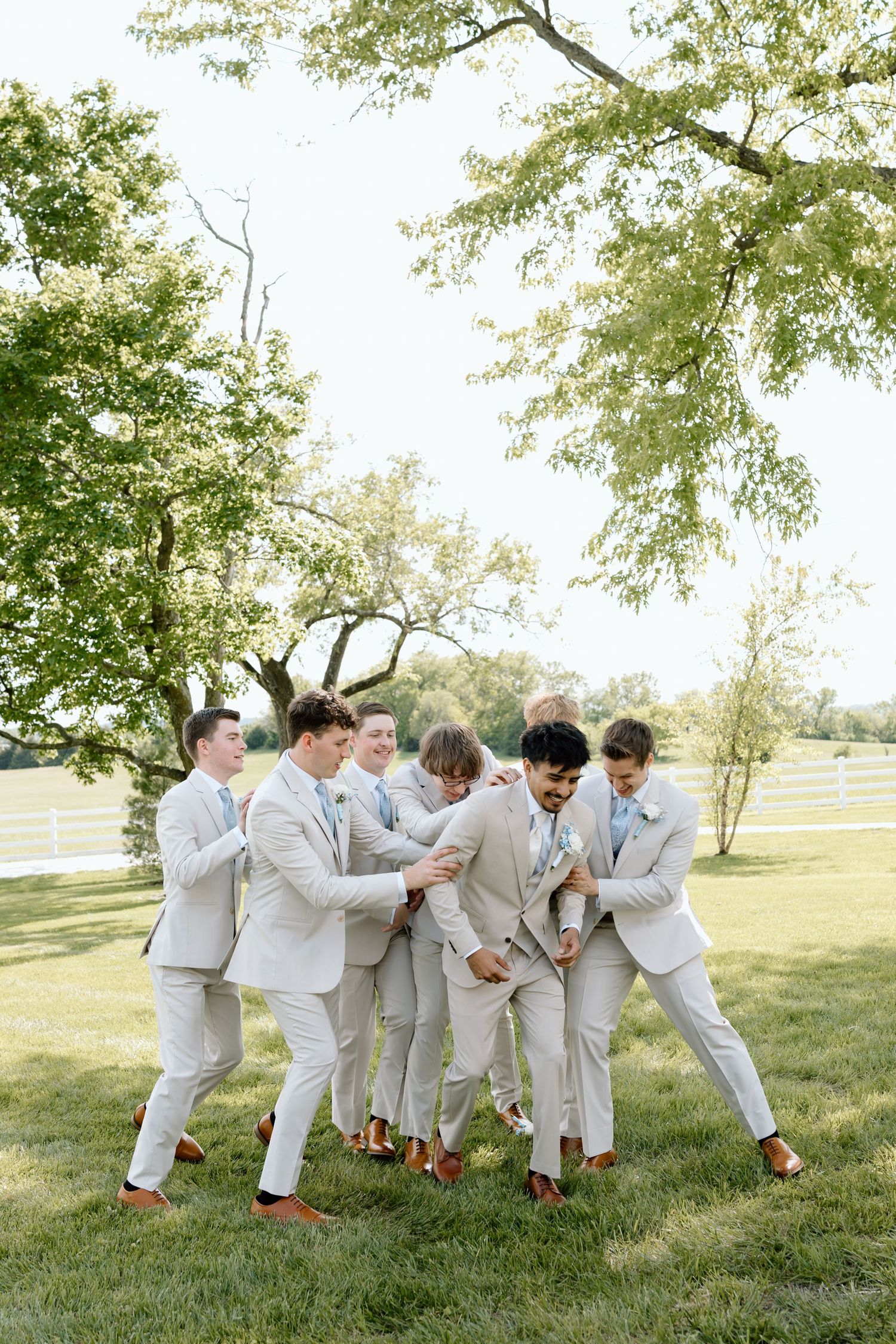 Groomsmen in tan suits playfully surround the groom outdoors on green lawn with trees in background at summer wedding.