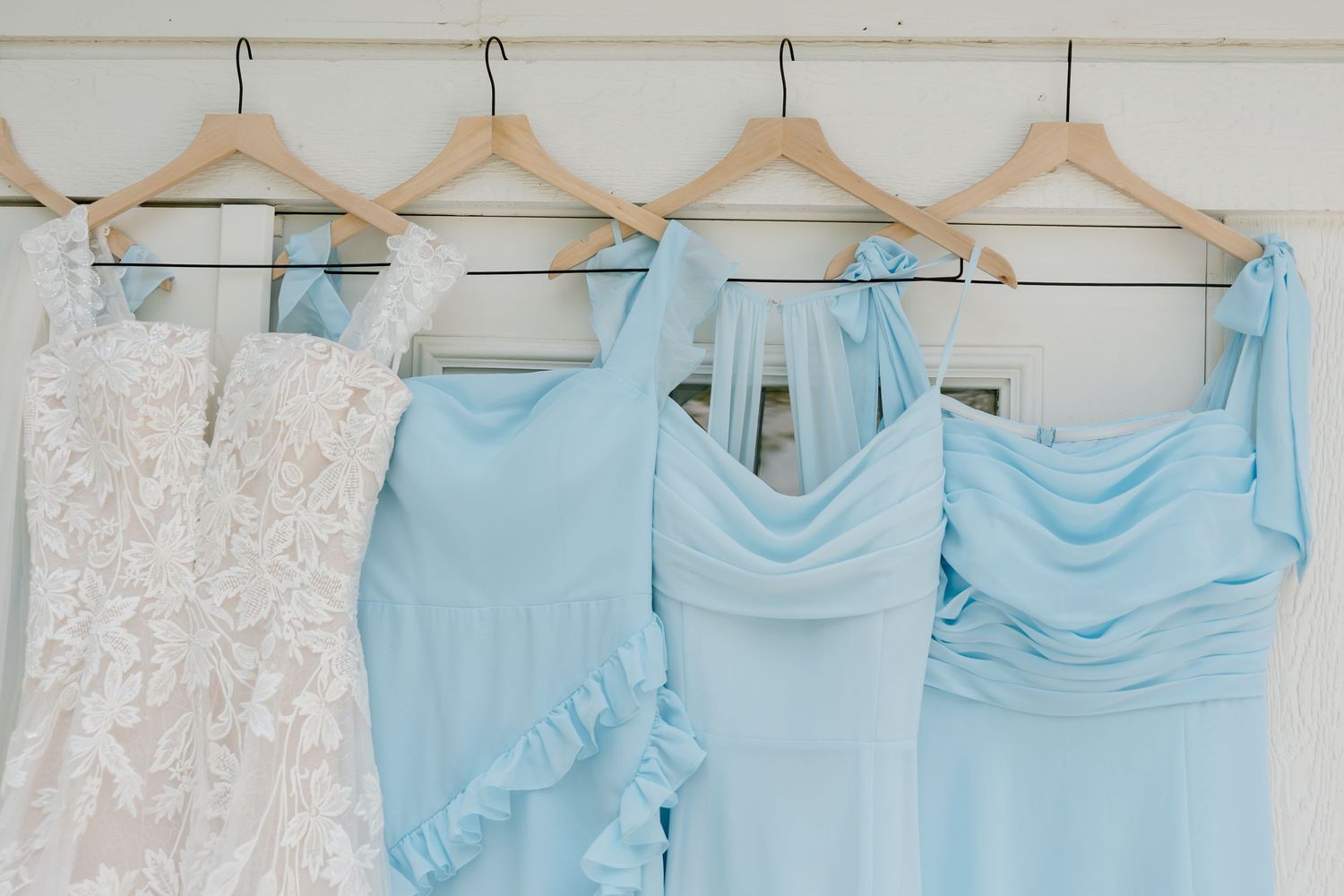 White lace wedding dress and light blue bridesmaid dresses hanging on wooden hangers against a white wall.
