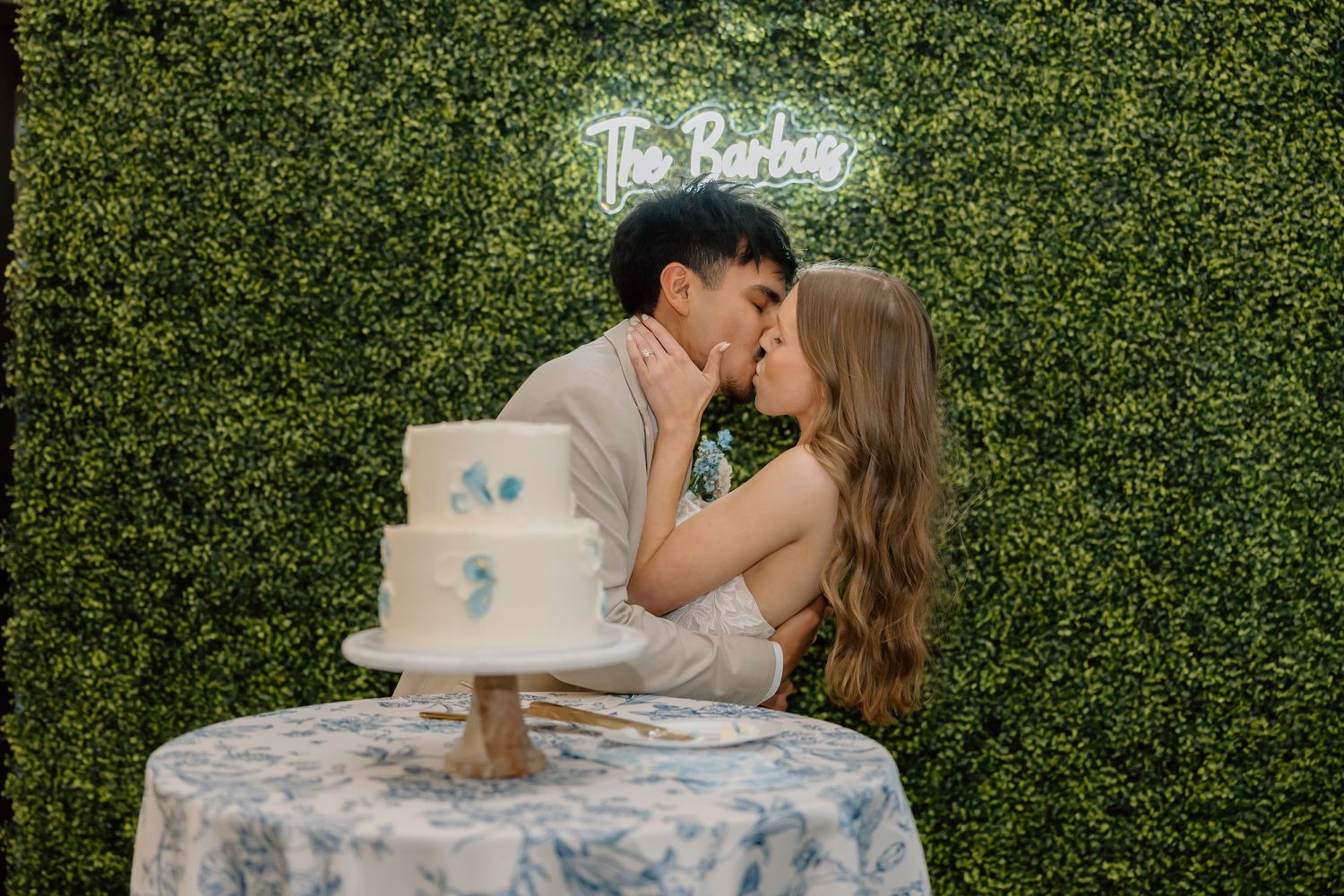 Newlyweds sharing a kiss at their wedding cake cutting in front of a green hedge wall with neon sign.