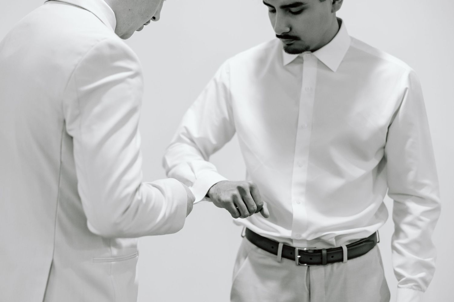 Two men in white dress shirts, one adjusting the other's belt in a black and white photo.