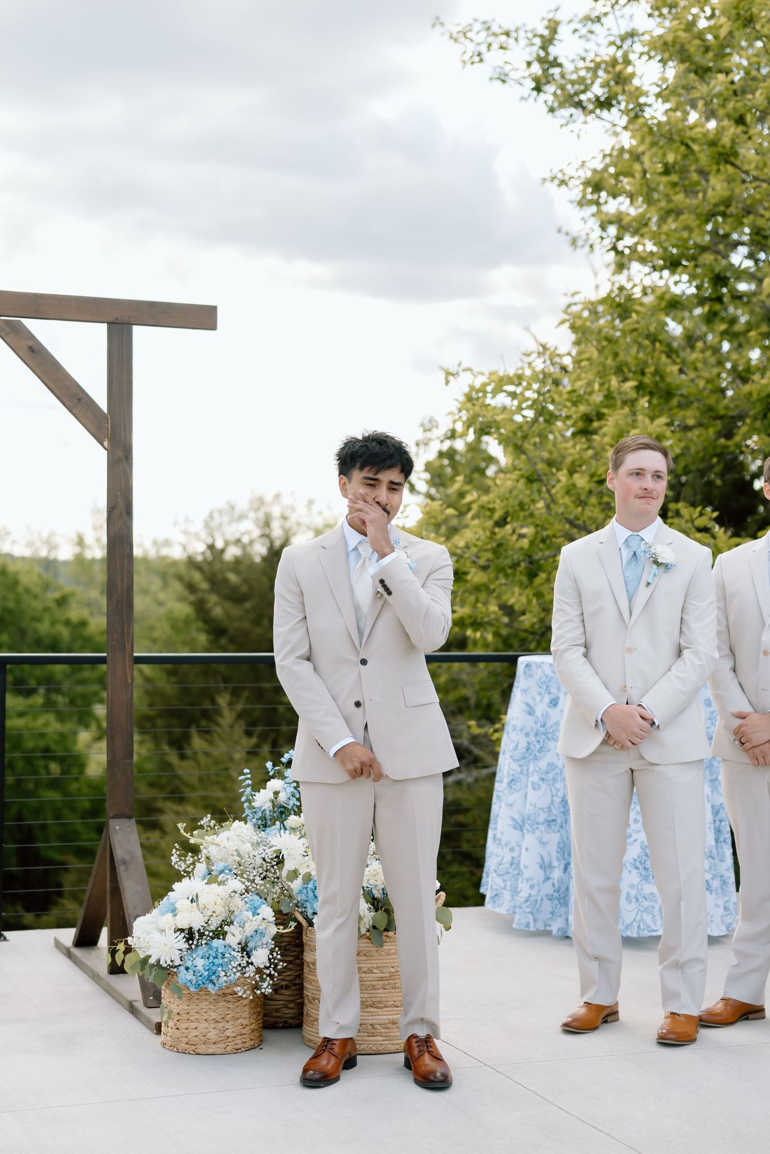 Emotional groom in beige suit covering mouth at outdoor wedding ceremony with blue floral arrangements and wooden arch.