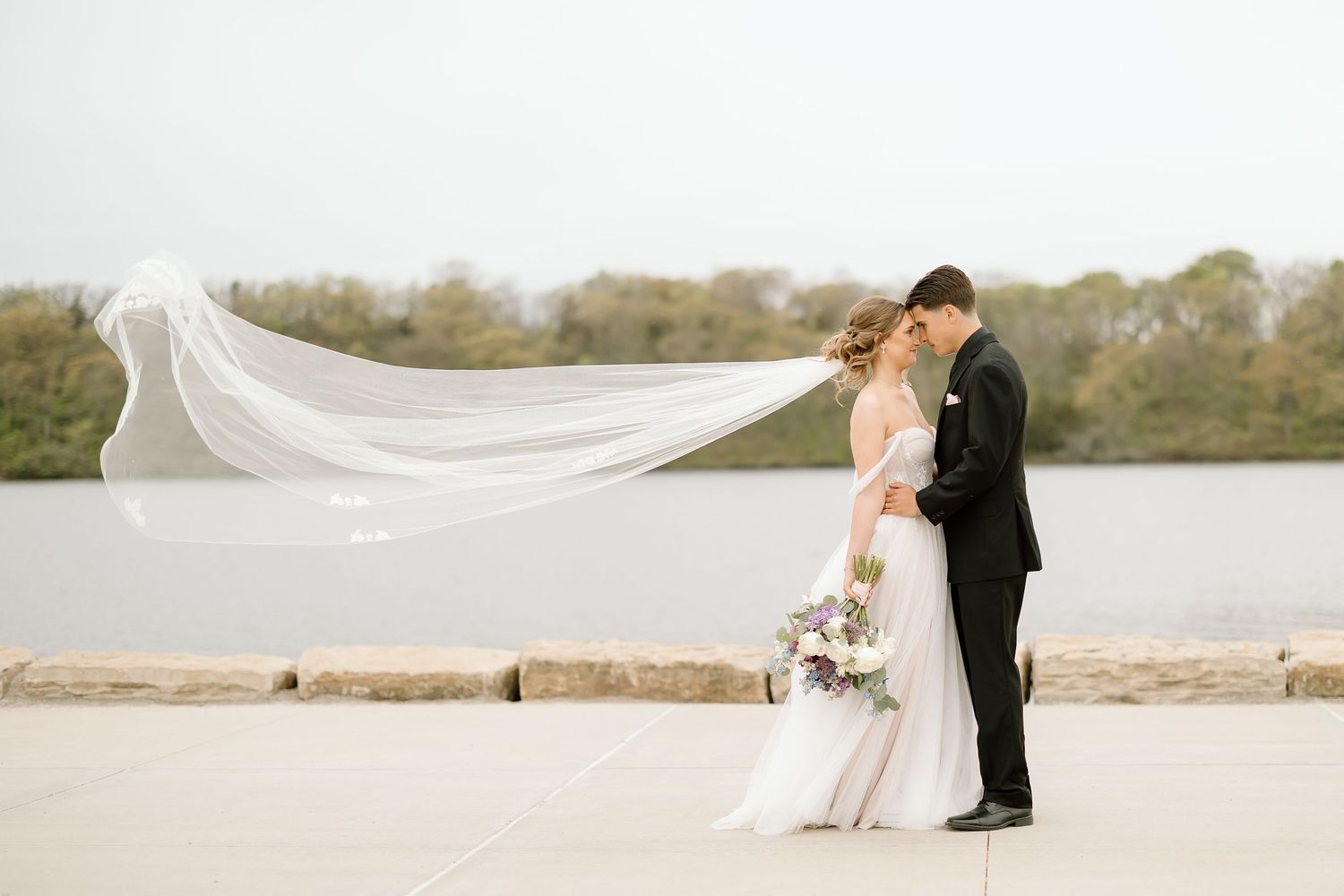 Wedding couple poses as veil flows dramatically in wind by waterfront.