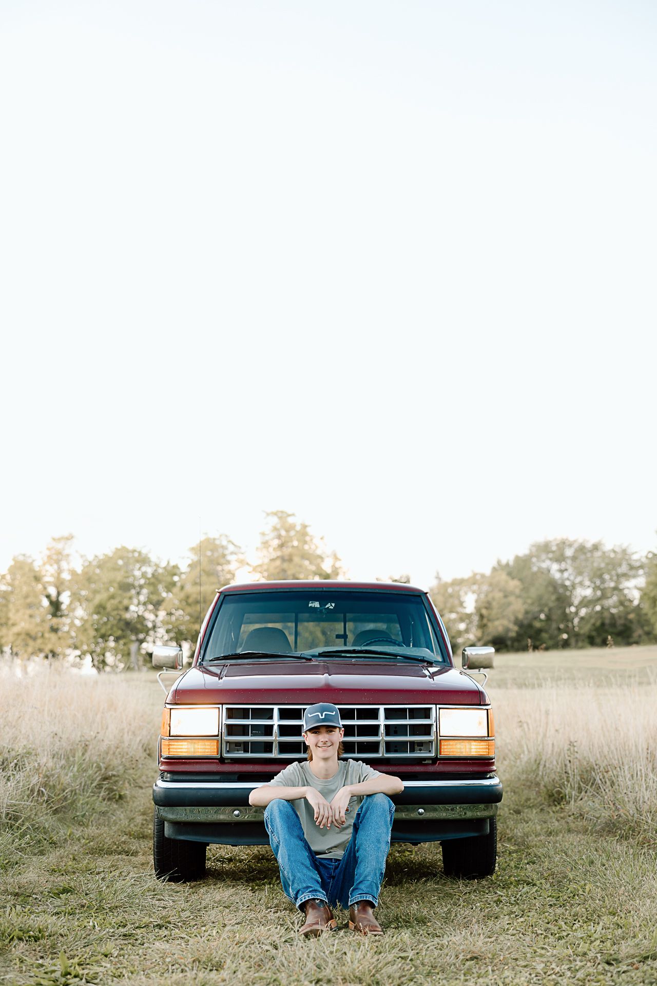 Front view of someone sitting on ground in front of a red Ford truck in tall grass.