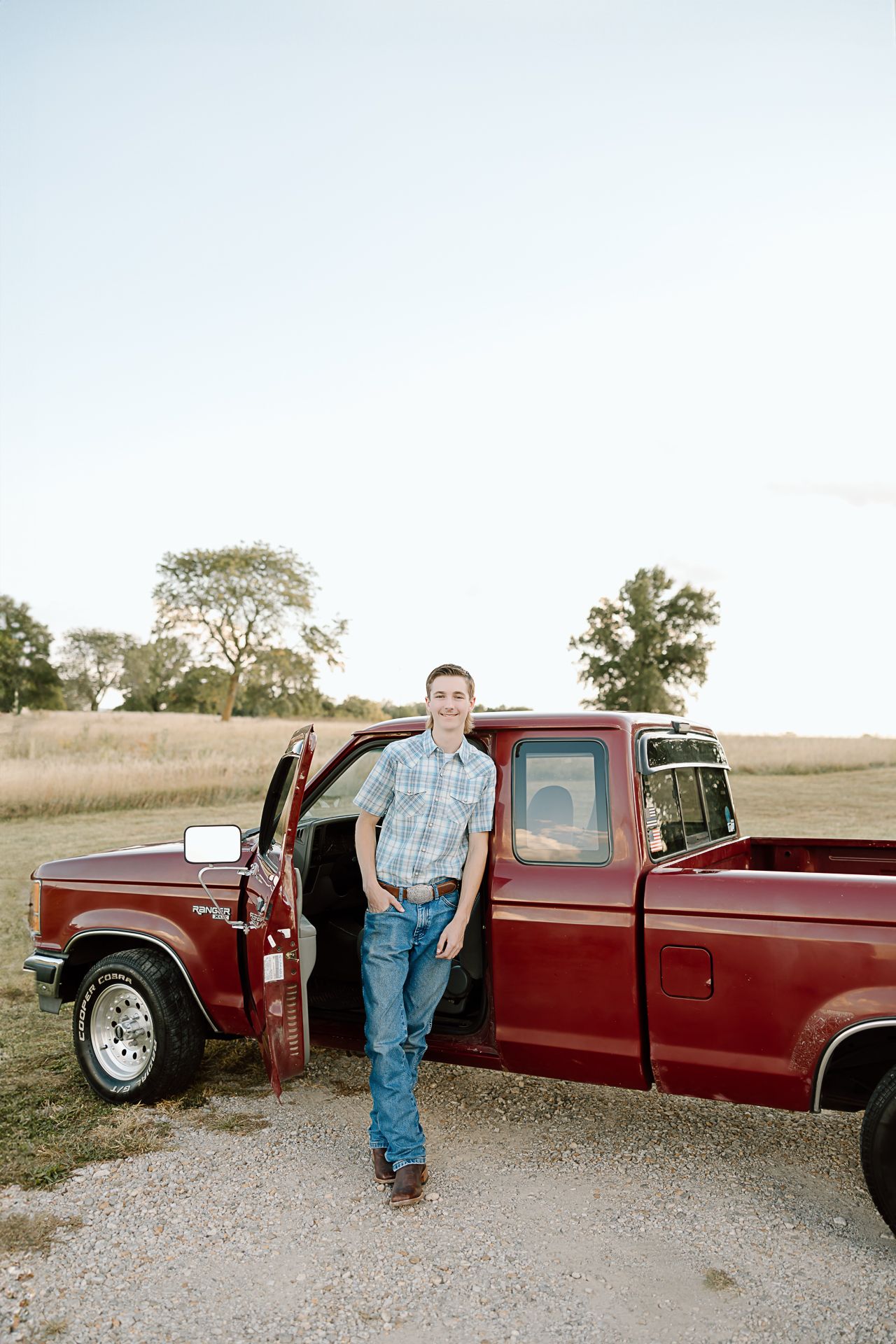 Someone posing casually against a classic red pickup truck in a countryside setting at golden hour.