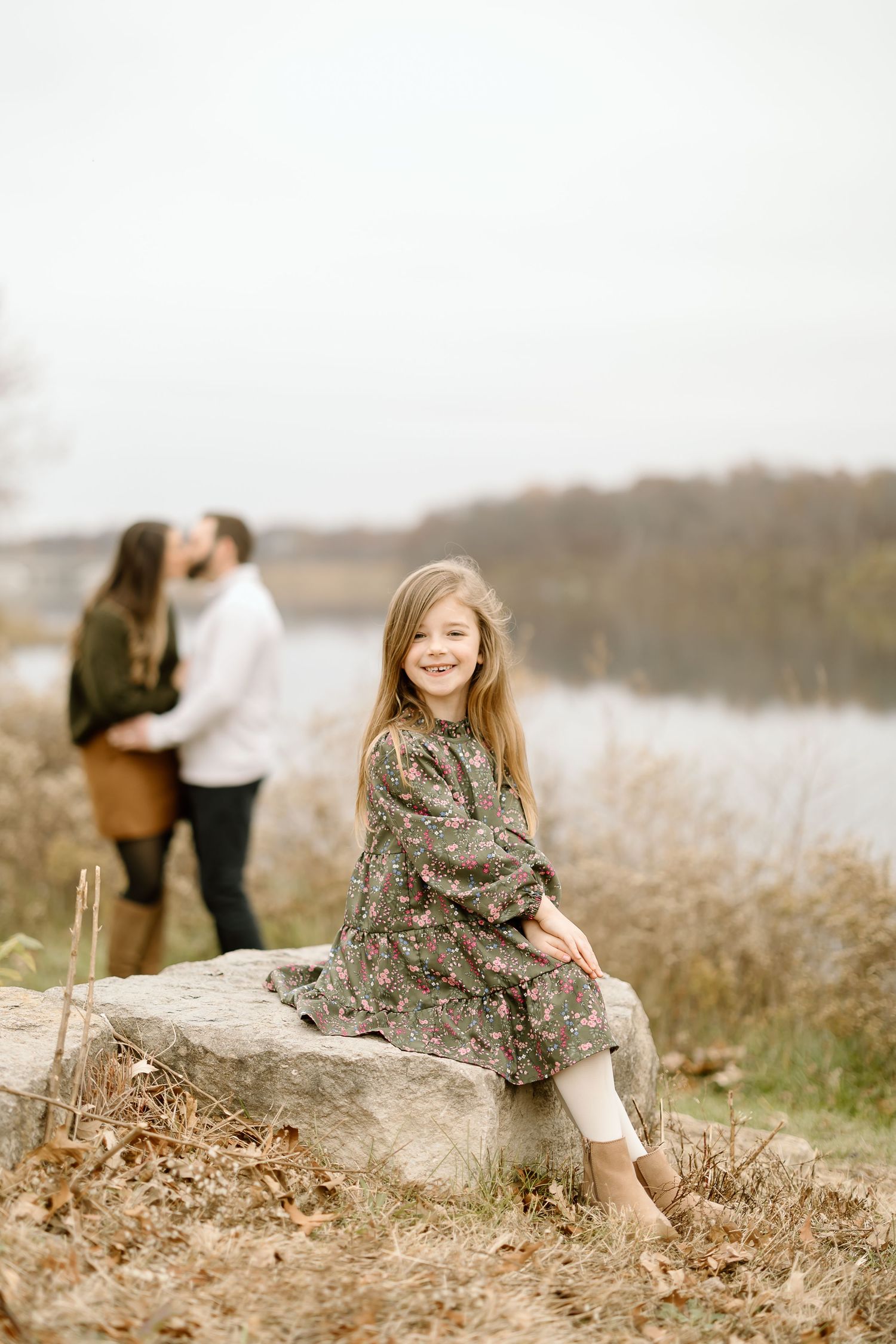 Child in floral dress sitting on rock by lake with two people in background.