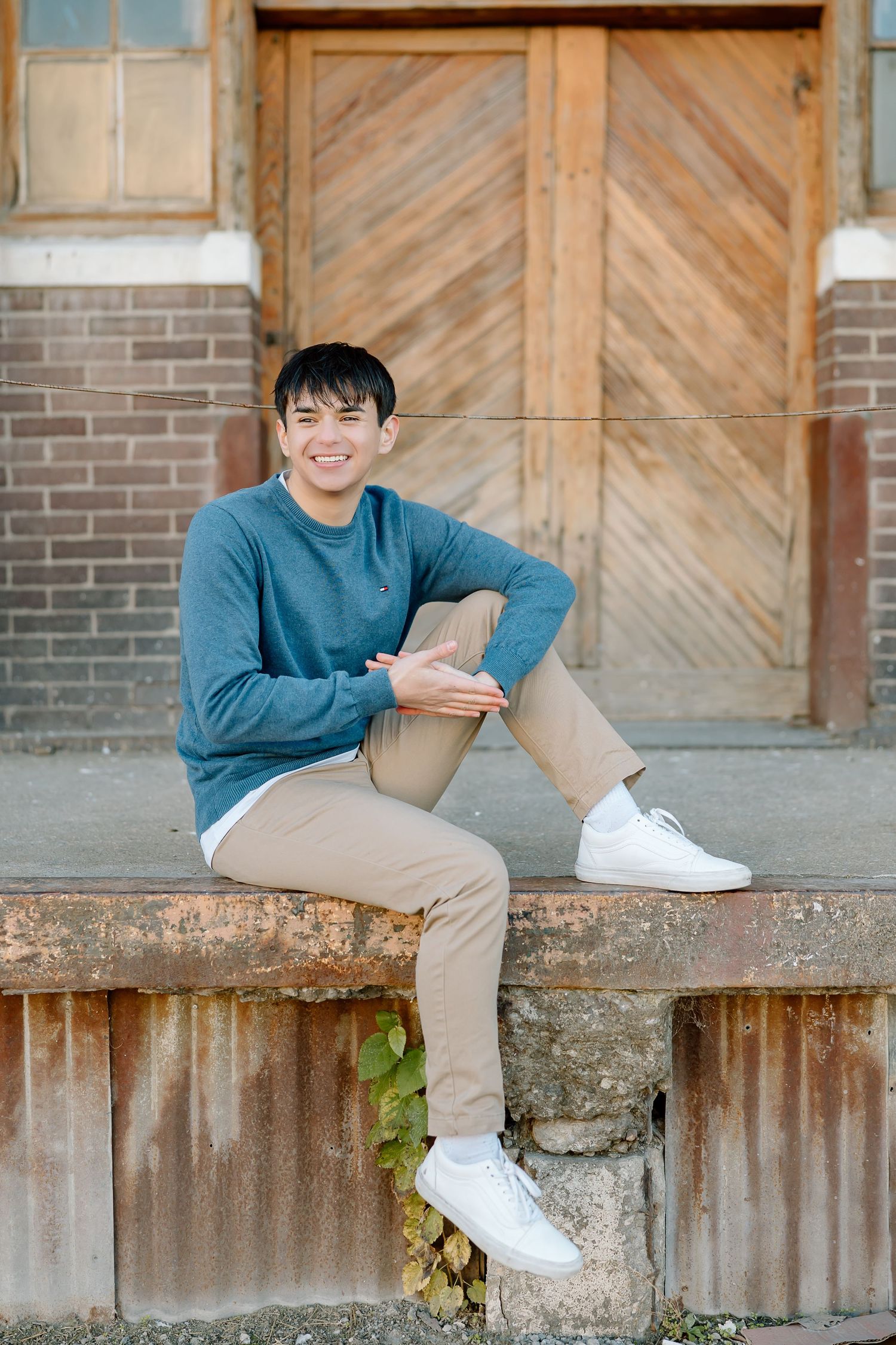 Relaxed portrait featuring person in blue sweater sitting on concrete ledge by vintage wooden door.
