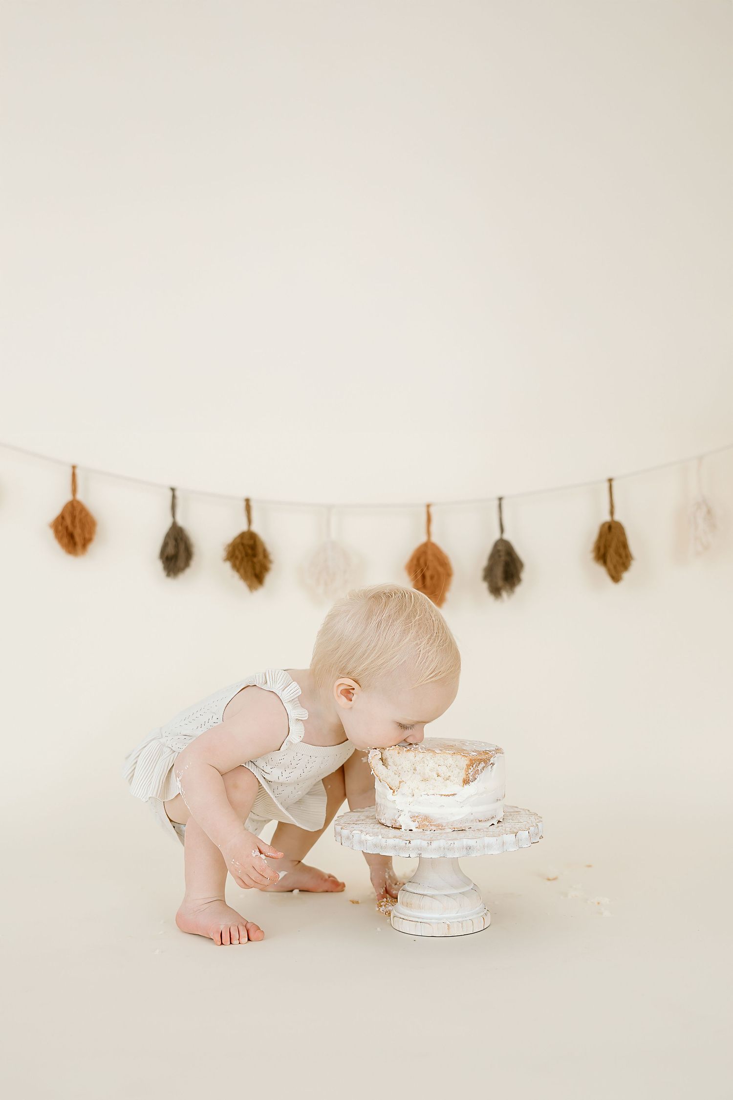 Three connected images show a baby in white crawling towards and tasting a white birthday cake with fall leaves hanging above.