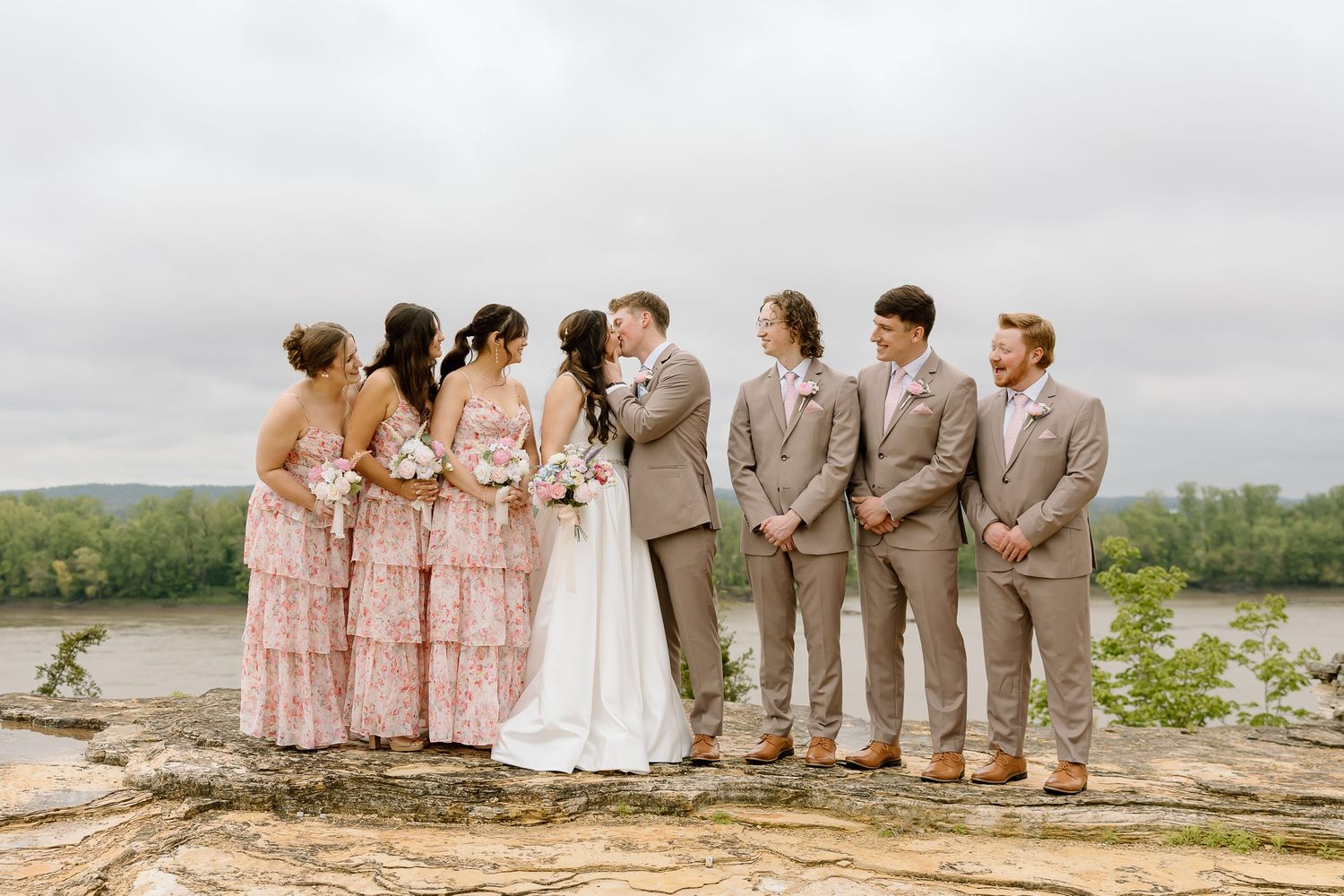 A wedding party in pink dresses and tan suits poses along a scenic rocky coastline on an overcast day.