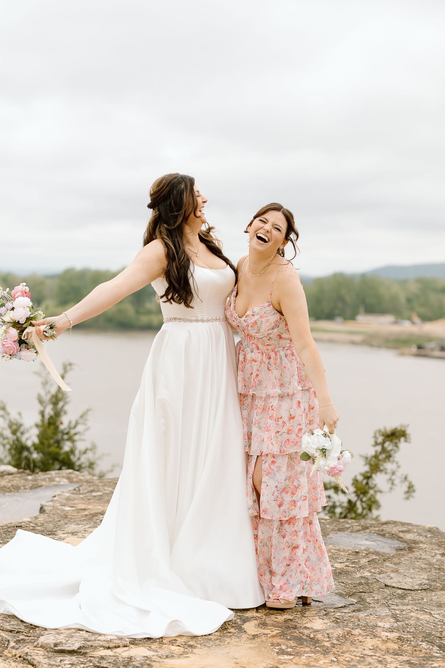 Two friends in white and pink dresses laugh together while sitting on rocks near a lake on a cloudy day.