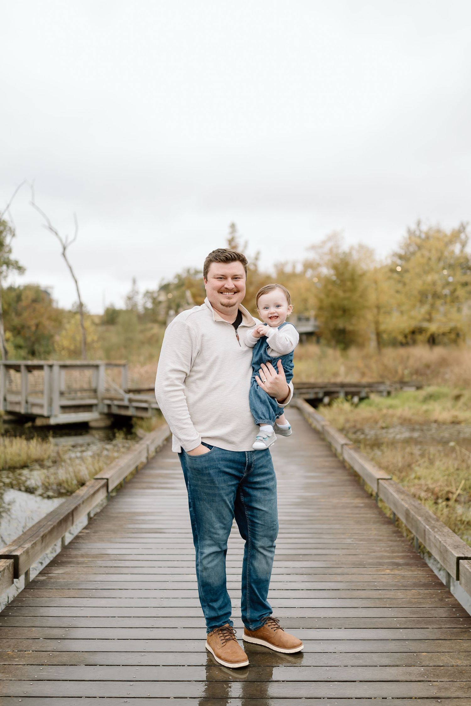 Two people standing on a wooden boardwalk surrounded by autumn foliage on an overcast day.