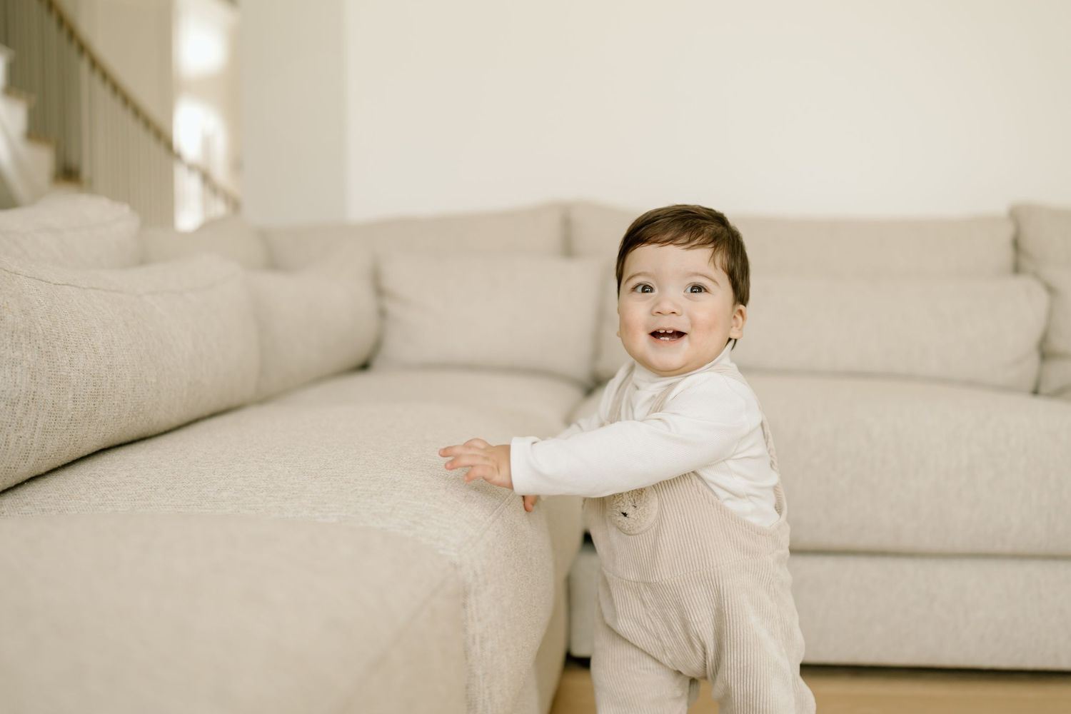 A happy toddler in a white sweater and beige pants stands next to a light gray couch in a bright room.
