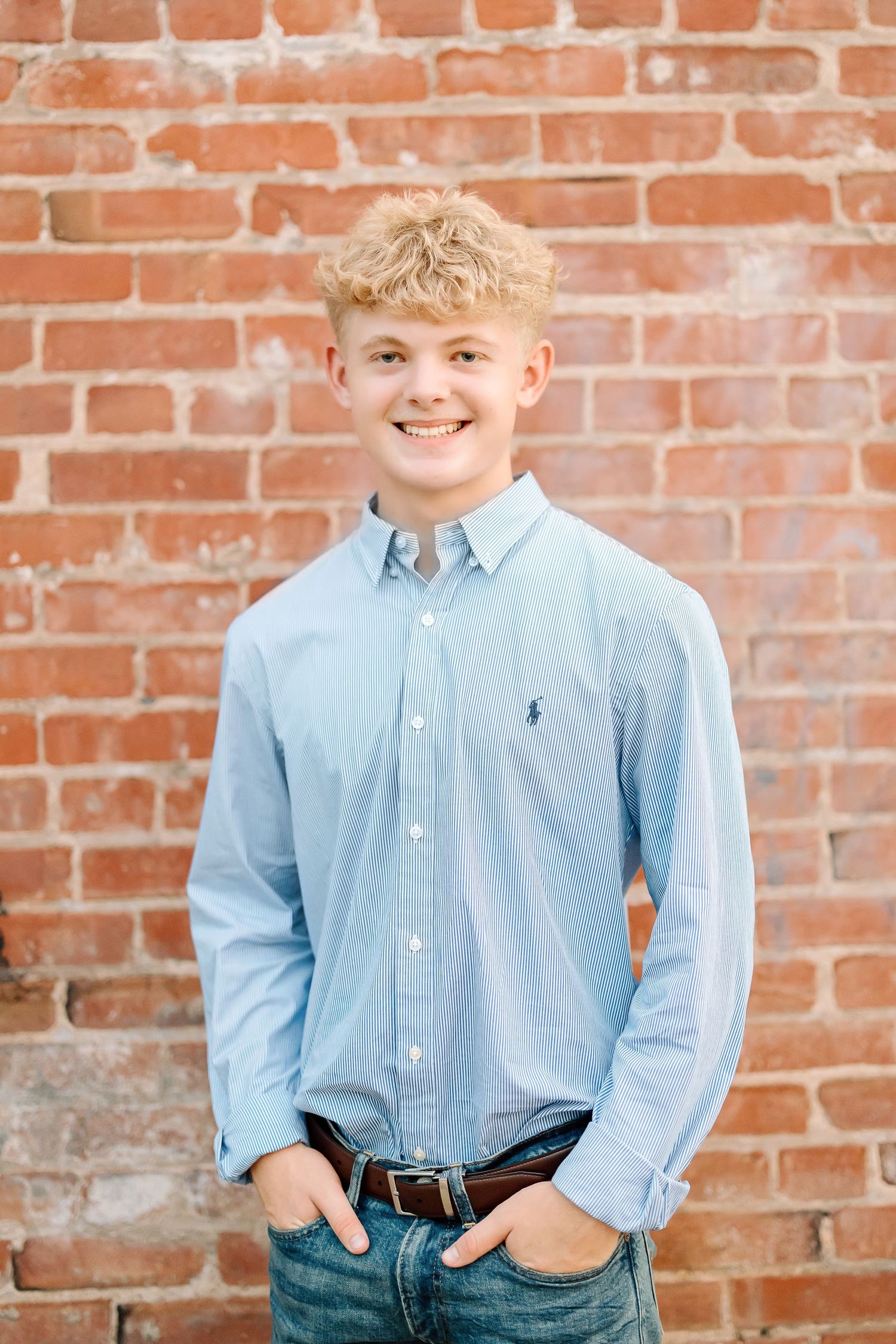 A person in a light blue button-down shirt and jeans poses casually against a red brick wall background.