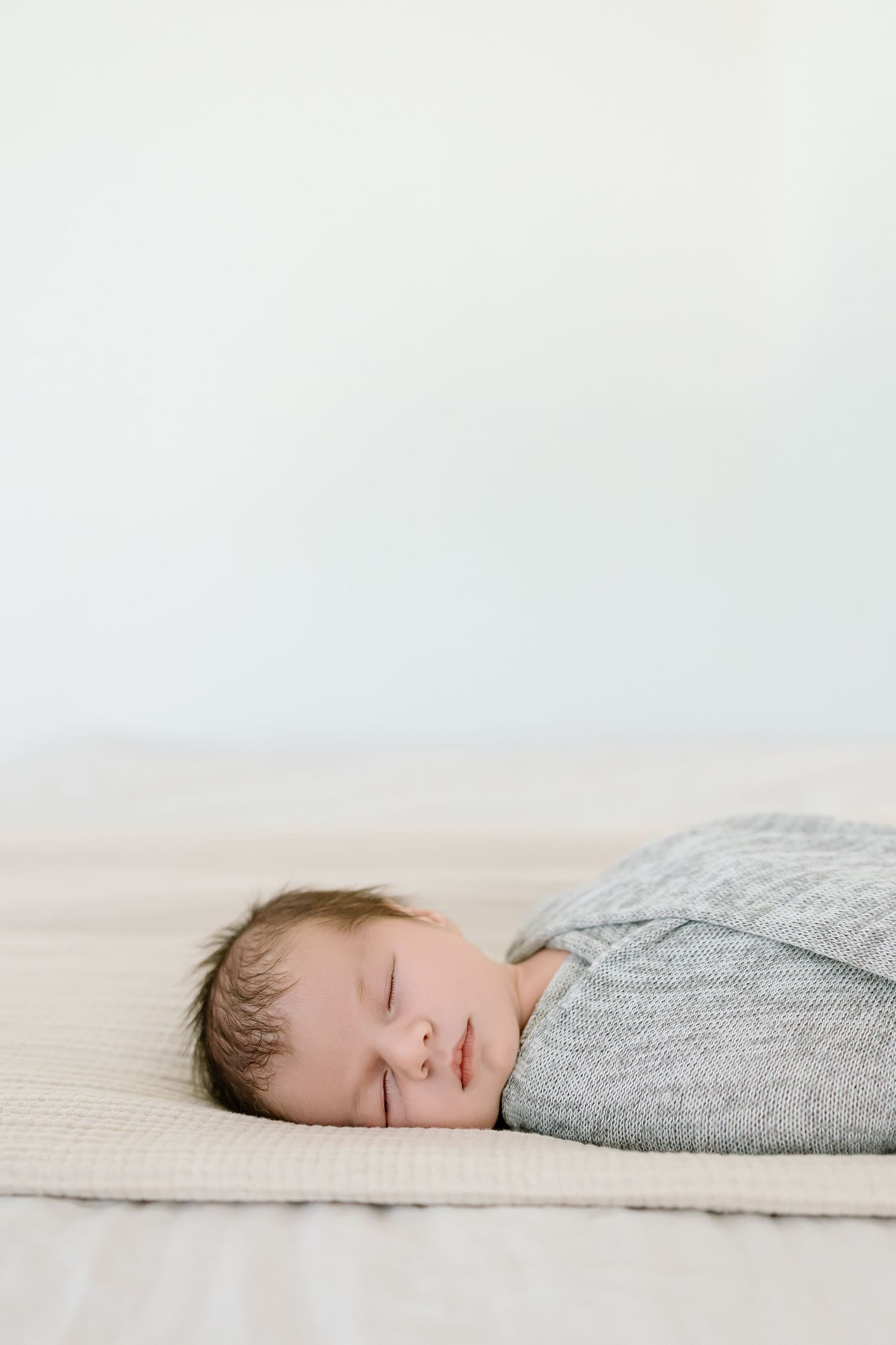 Peaceful sleeping newborn baby wearing grey swaddle lying on cream colored surface.